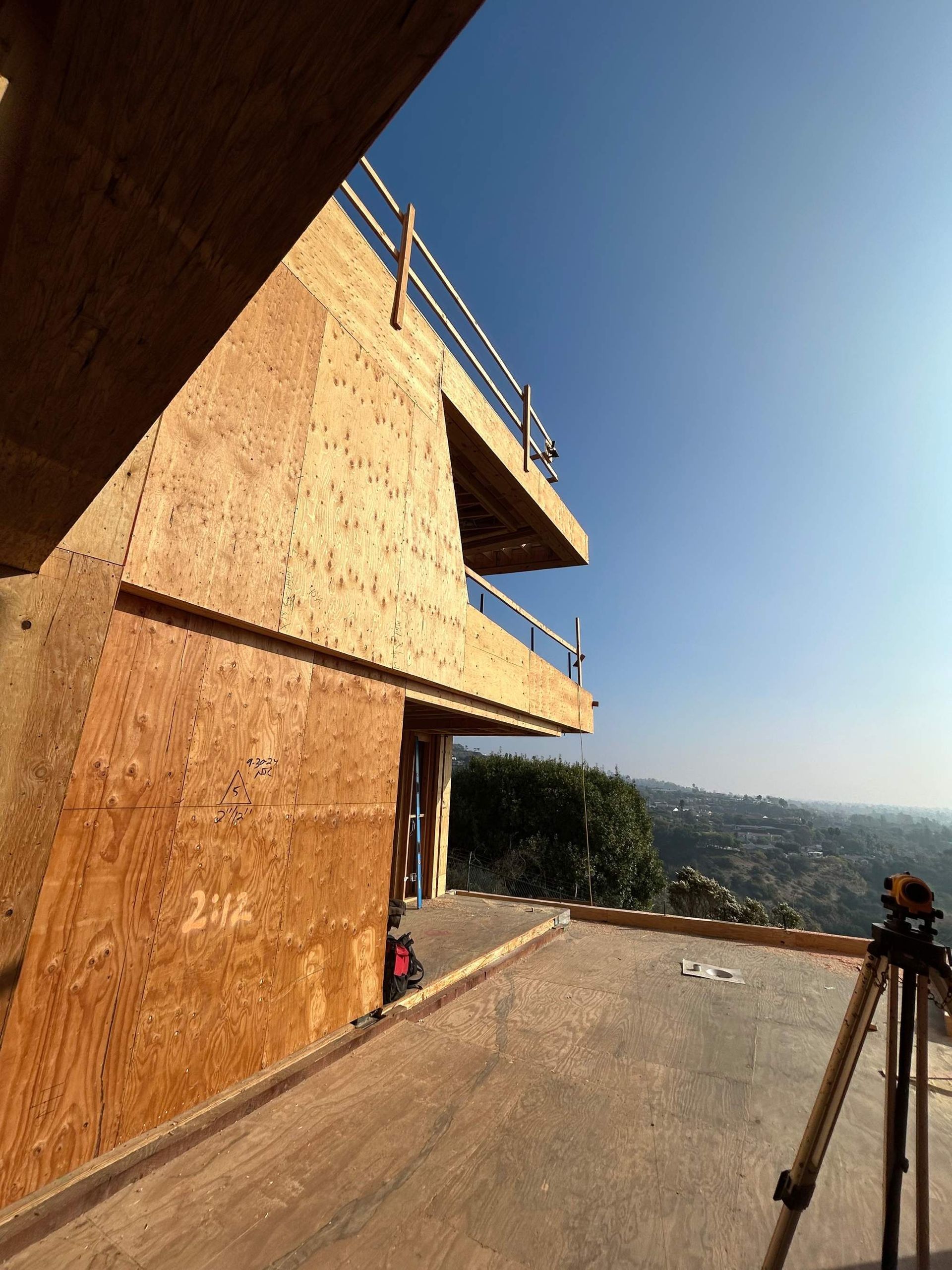 Wooden building under construction with plywood walls and a tripod on a rooftop, overlooking a hazy landscape.