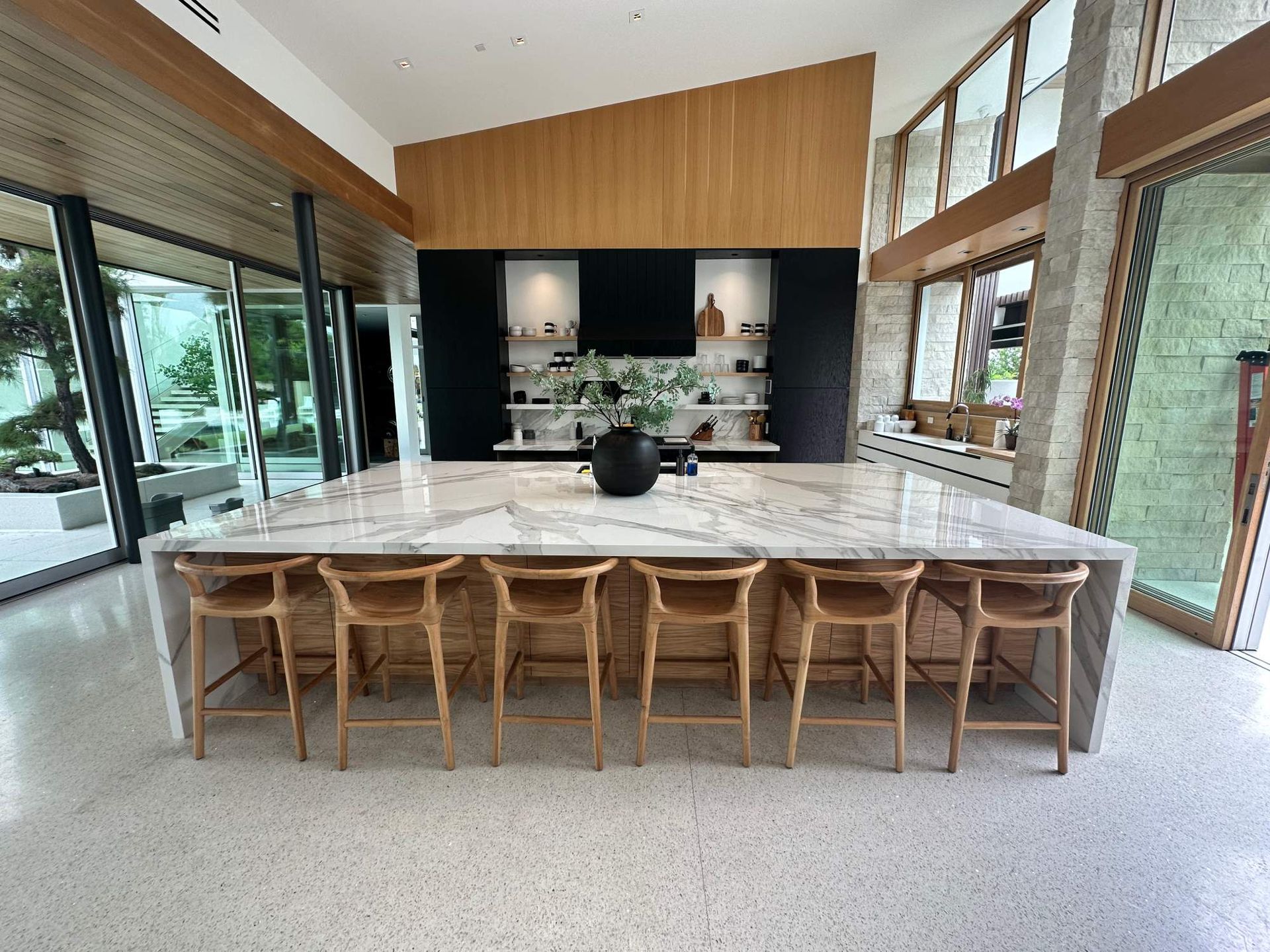 Modern kitchen with large marble island, wooden stools, and floor-to-ceiling windows facing greenery