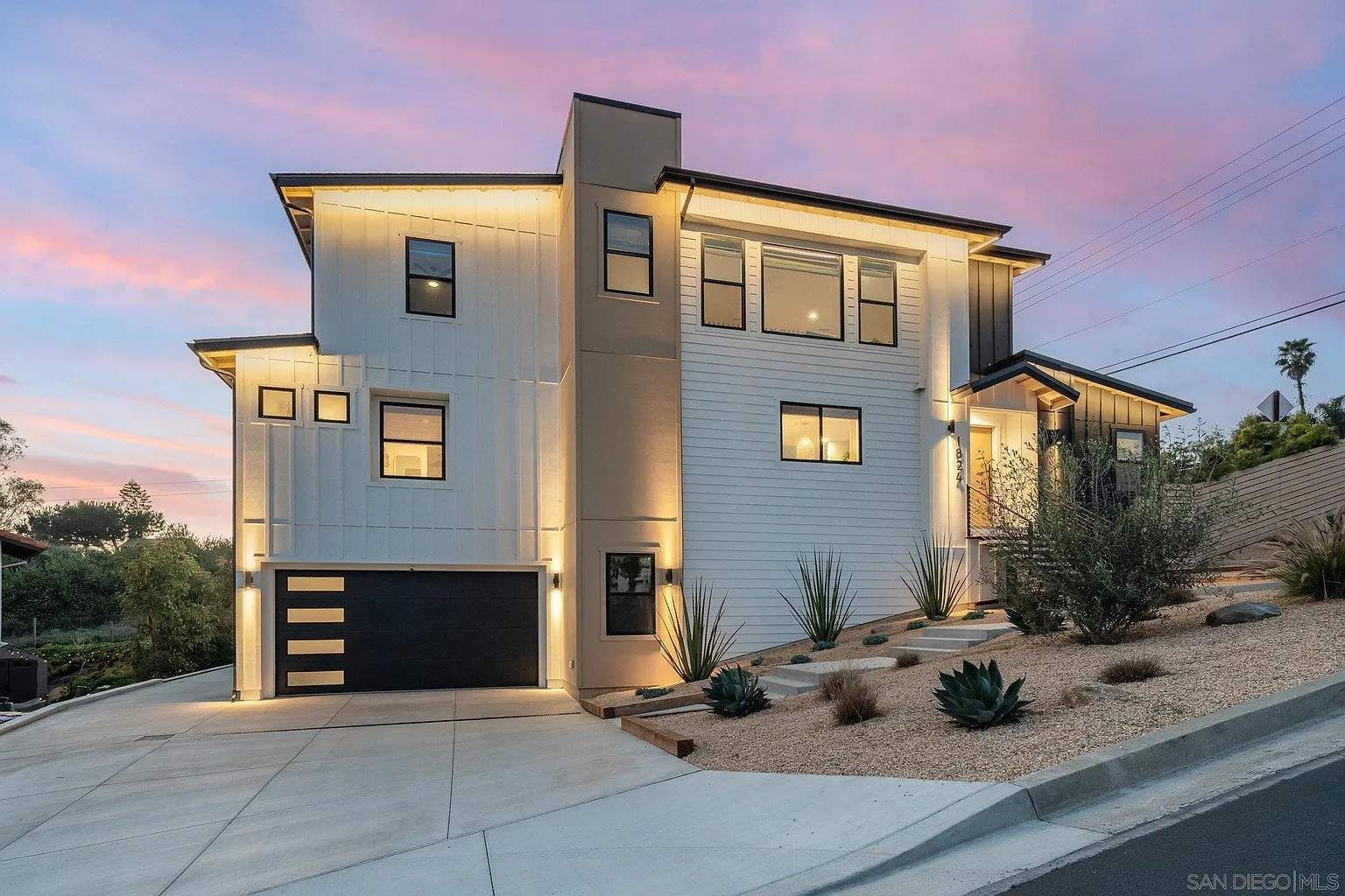 Modern two-story hillside house at dusk with lit windows and a pink sky