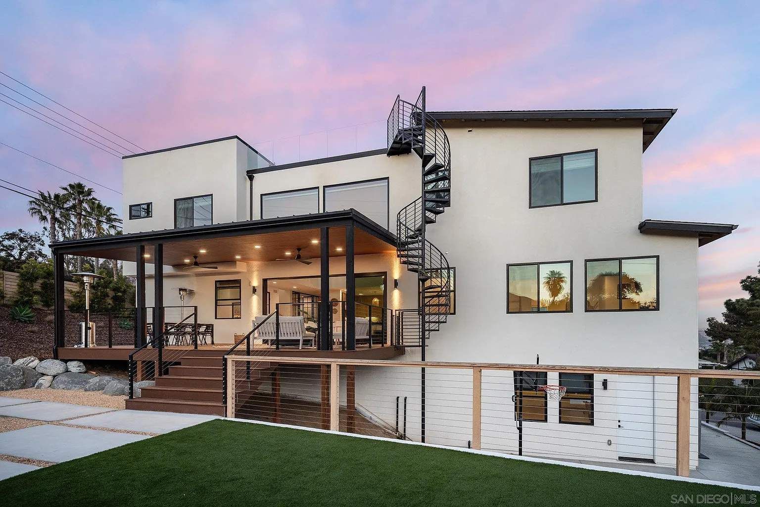 Modern two-story house with a lit patio, spiral staircase, and landscaped lawn at dusk