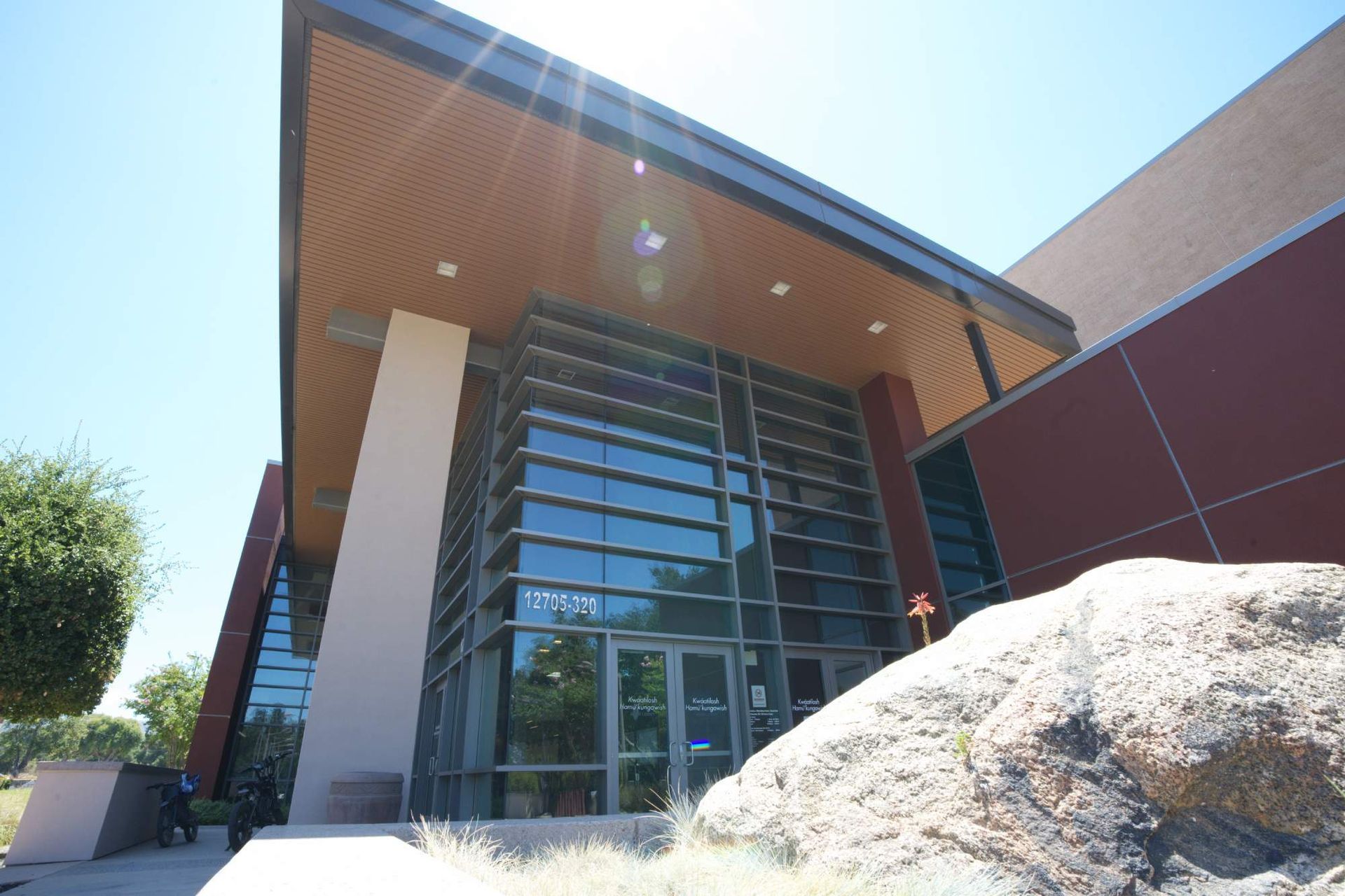 Modern building entrance with glass doors, wood overhang, and a large rock in the foreground