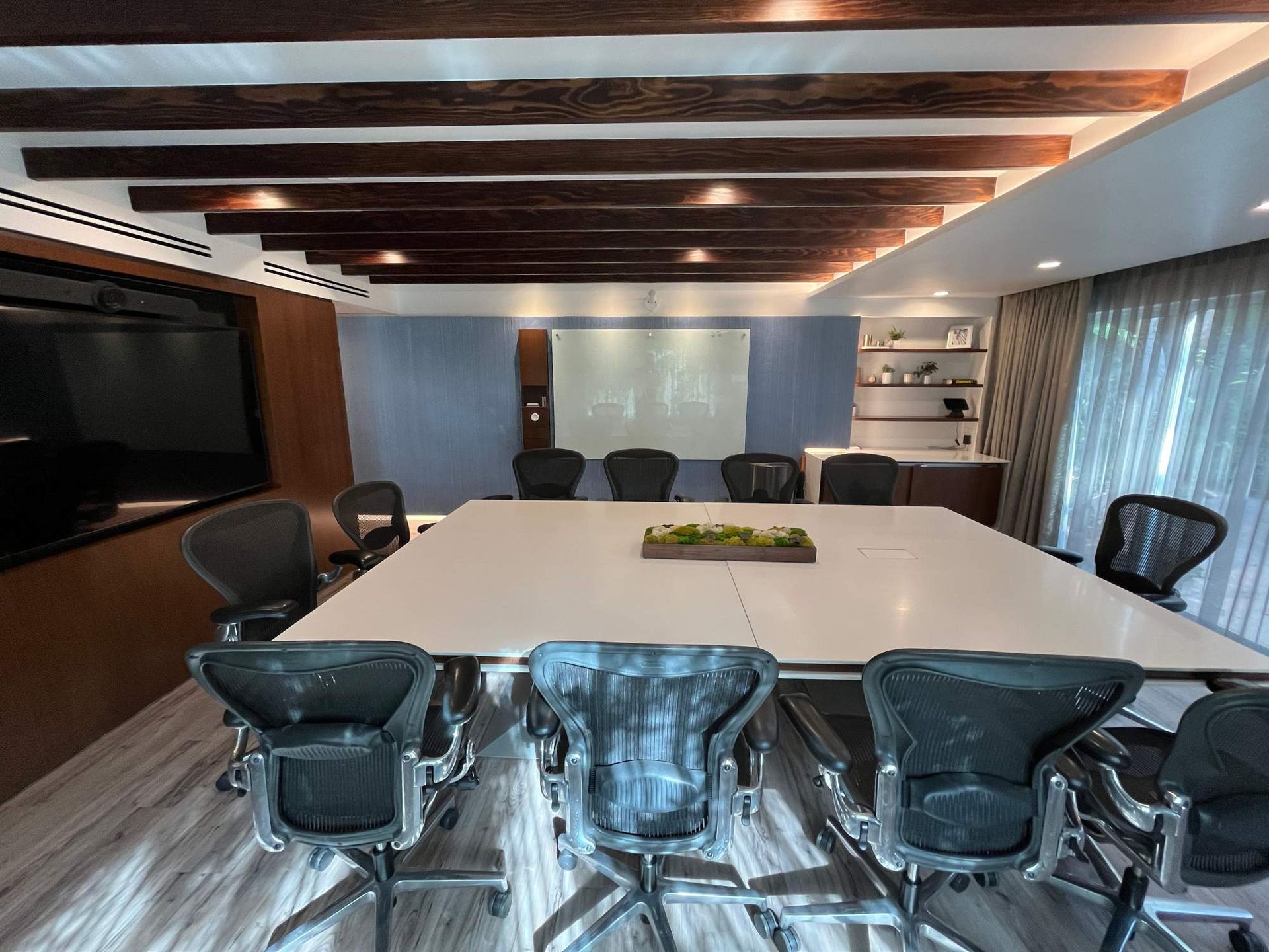 Modern conference room with white table, black chairs, wood ceiling beams, and large screen at the front