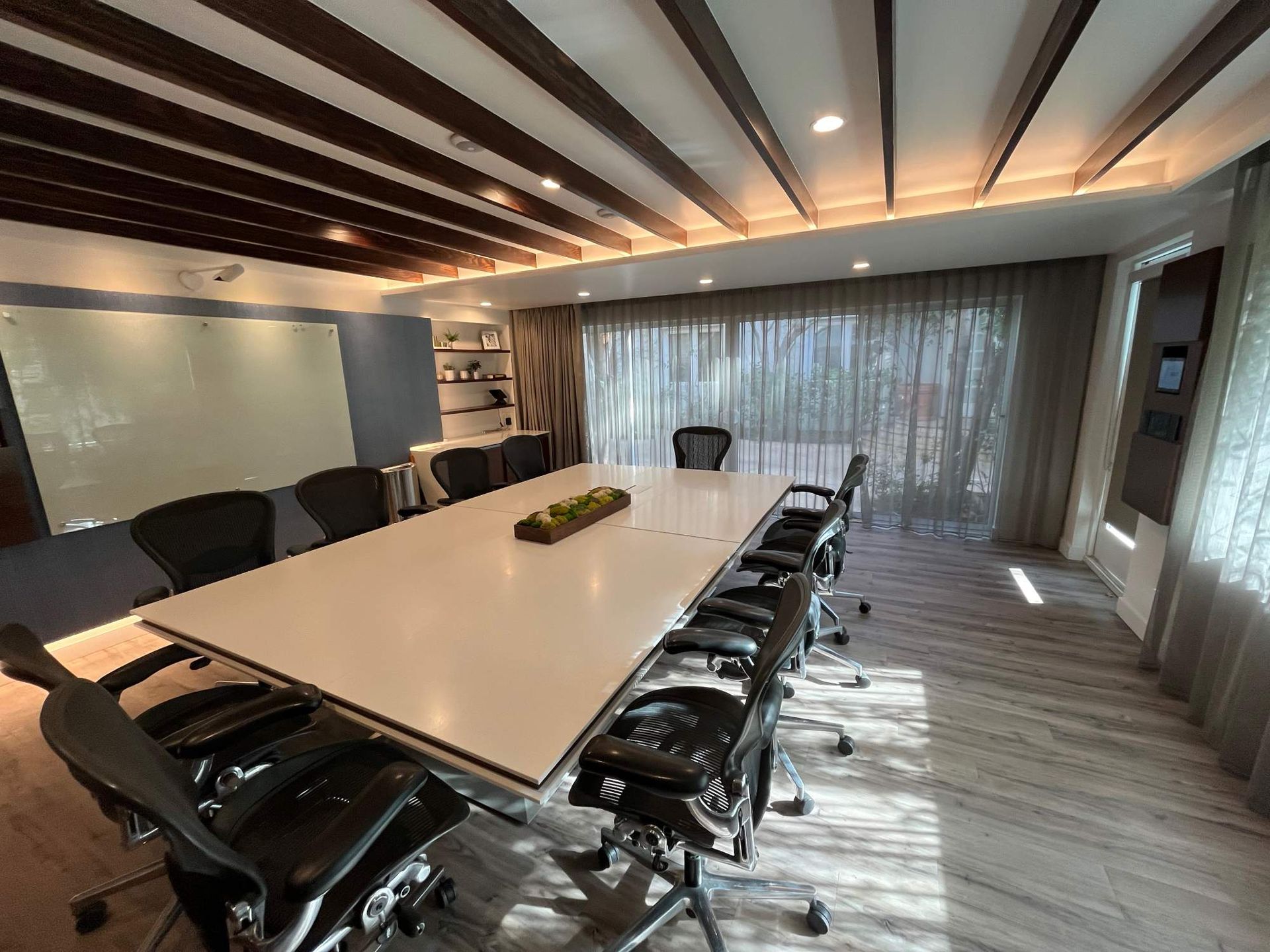 Modern conference room with long white table, black chairs, wood ceiling, and large windows with sheer curtains
