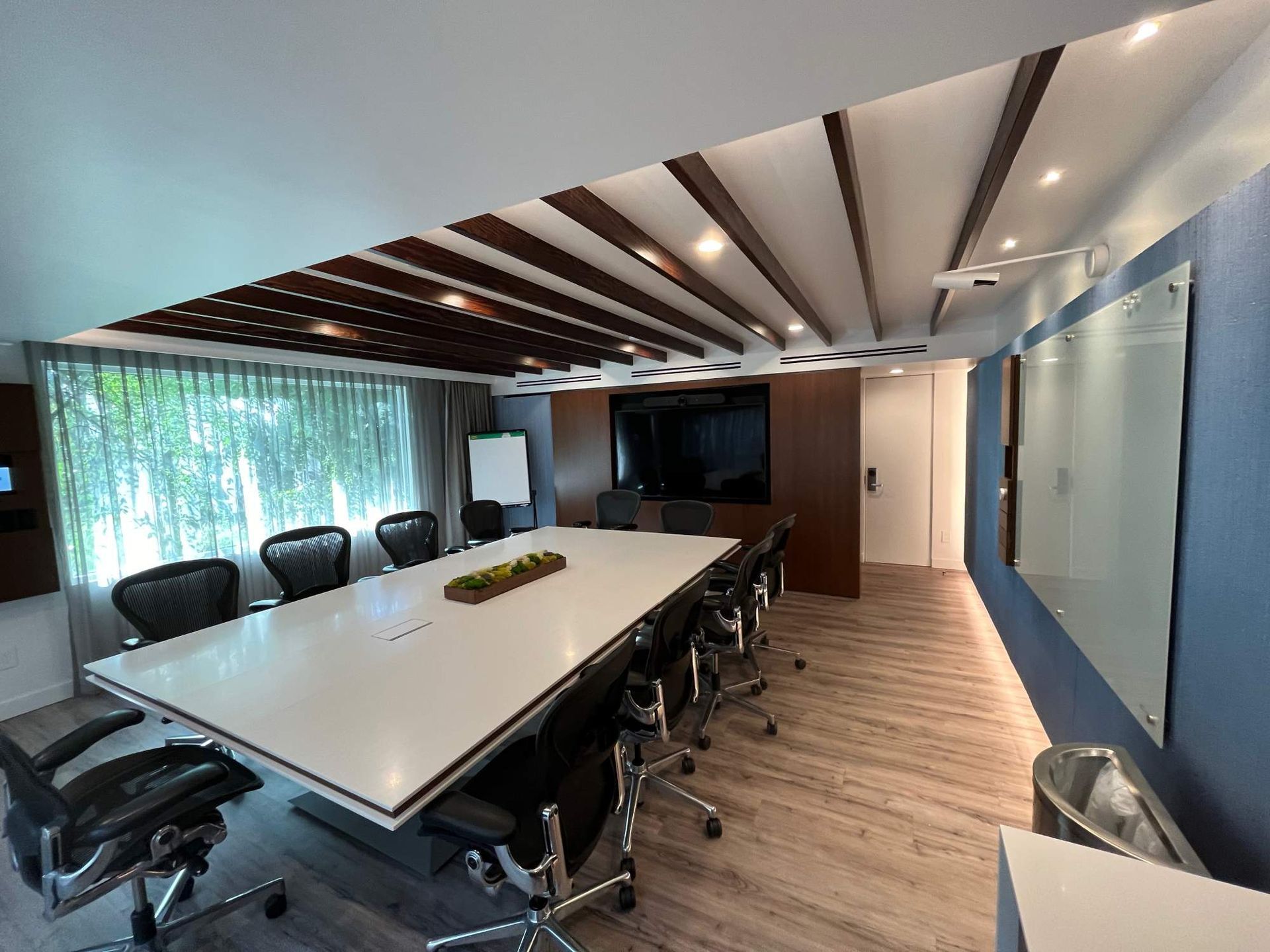 Modern conference room with long table, black chairs, wood ceiling accents, and large wall-mounted display