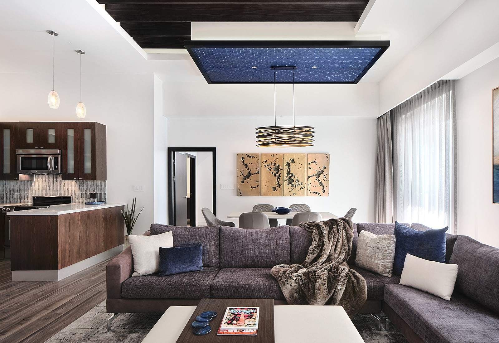 Modern open-concept living room with dark sectional sofa, coffee table, kitchen bar, and blue-lit recessed ceiling panel