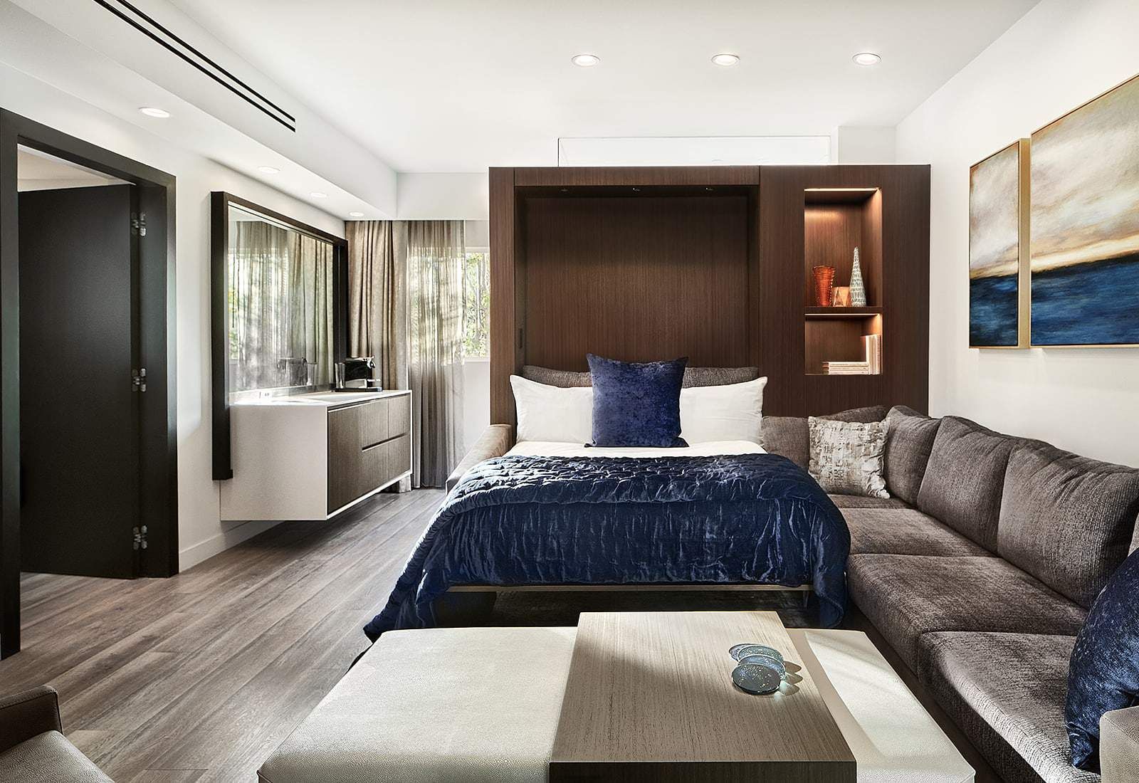 Modern hotel bedroom with gray sofa, navy bed, wooden alcove, and window seating area.