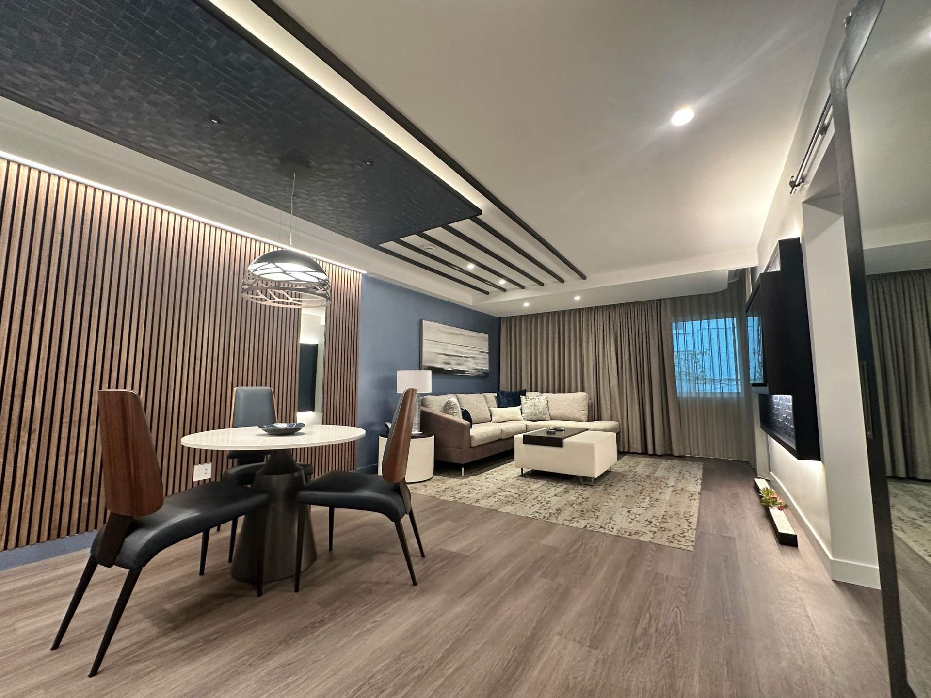 Modern living room with wood-paneled wall, dining table, beige sofa, and large window with curtains