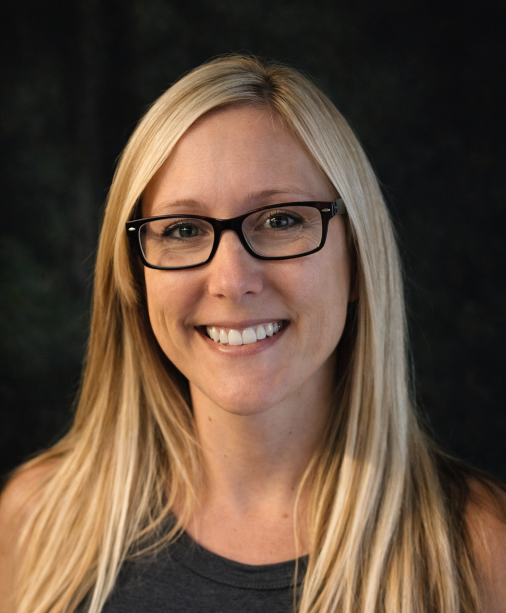 Smiling blonde person wearing glasses and a dark top against a dark background
