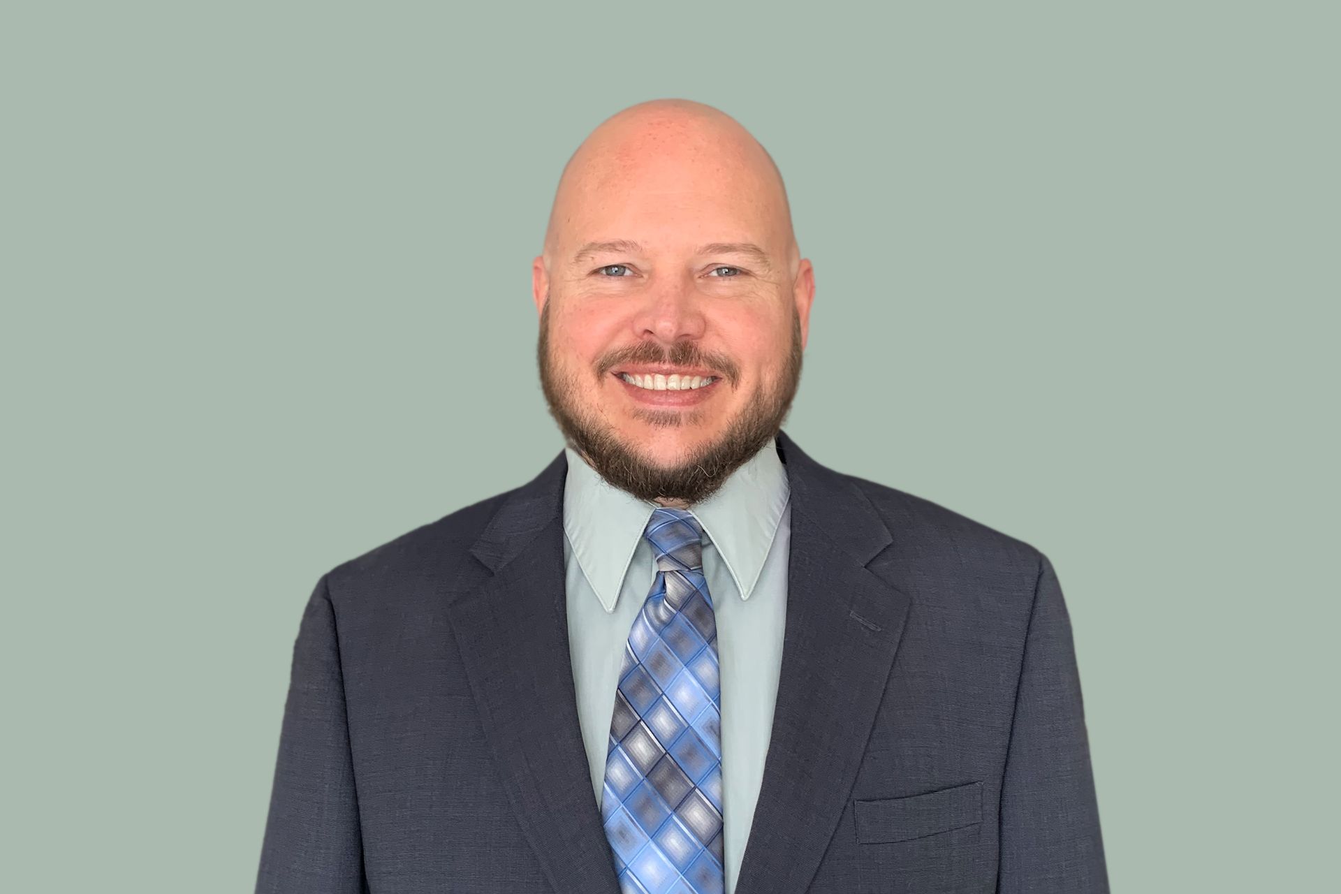 A smiling person with a beard, wearing a blue suit, light-colored dress shirt, and a blue plaid tie against a green backdrop.