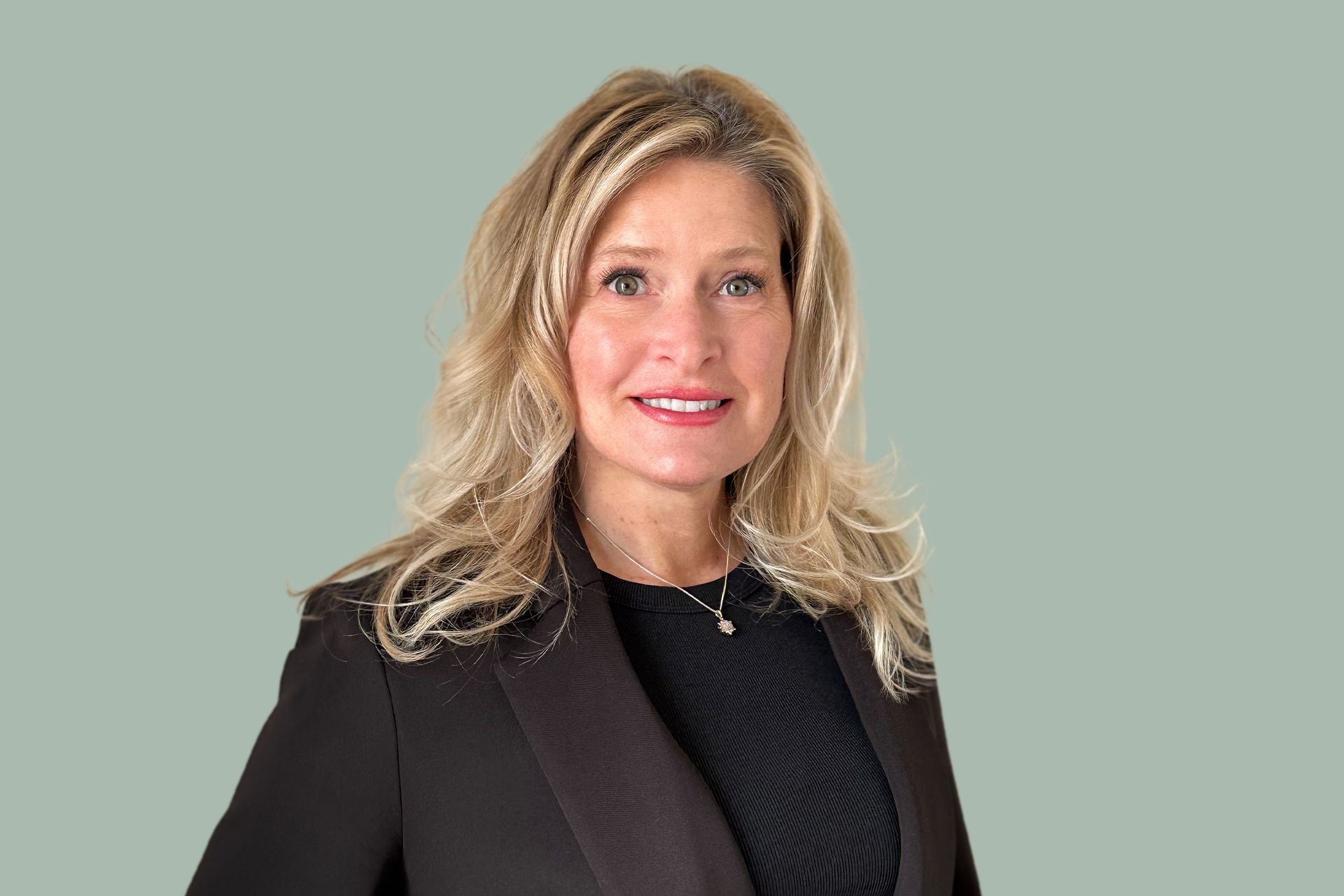 Kevin Hoskins A smiling woman with blonde hair, wearing a black blazer over a black top, poses against a light green background.