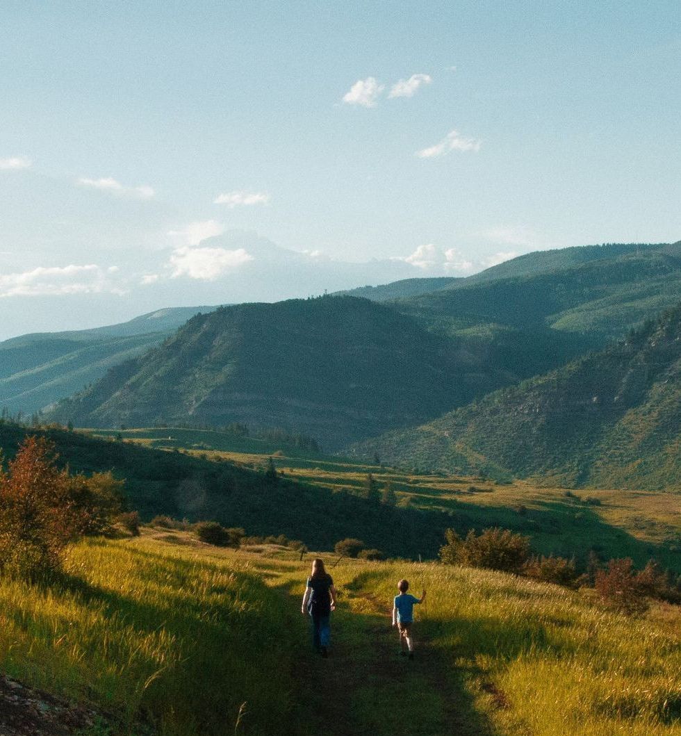 Two people walk on a dirt path through a grassy mountain meadow under a bright sky.