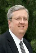 A professional headshot of a person wearing a dark suit, white shirt, patterned tie, and glasses, set against greenery.