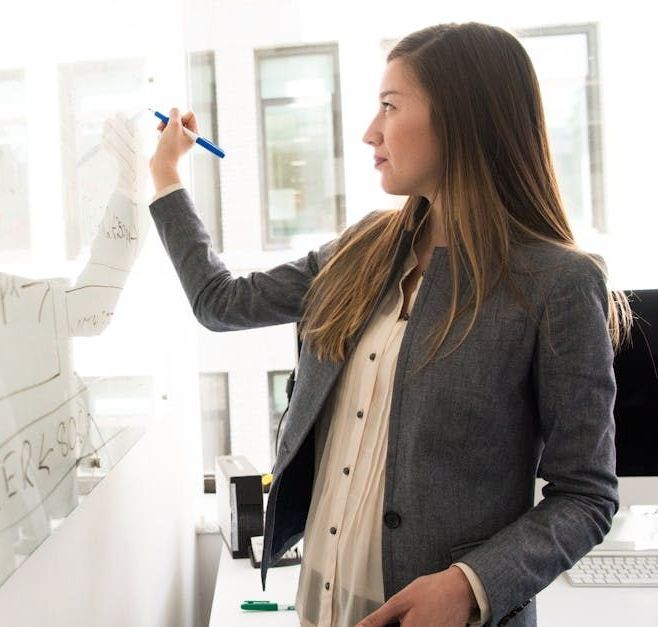 A person in a gray blazer writes on a glass board in a bright, modern office space.