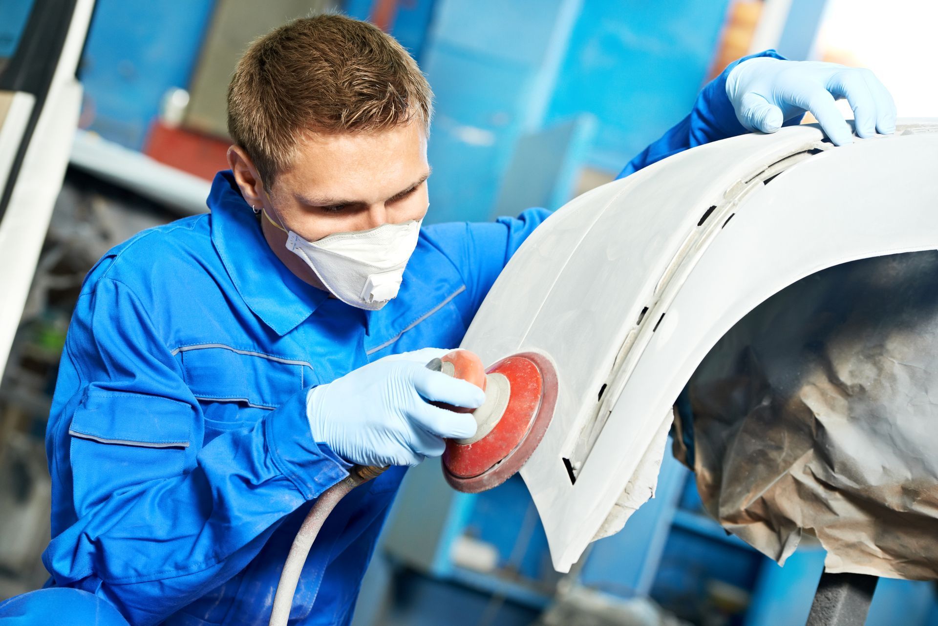 A man wearing a mask and gloves is sanding a car fender.