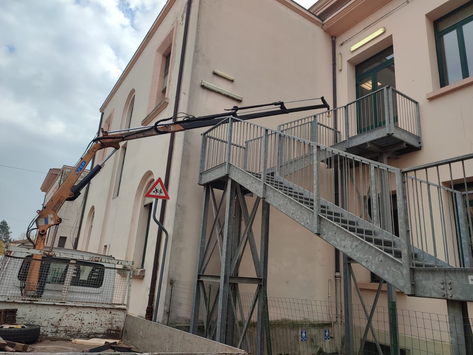 Una gru da cantiere parcheggiata accanto a un edificio rosa pallido con una scala esterna in metallo.