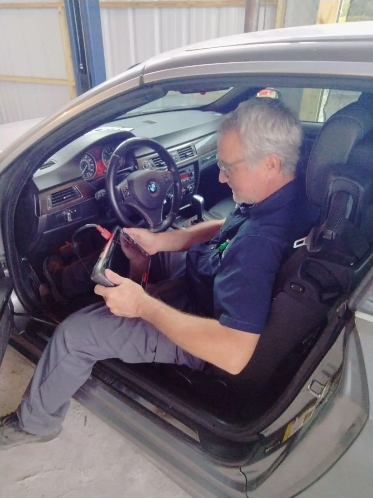 Man in a car, using a tablet. Interior shot. Gray car, open door. He wears a blue shirt and glasses.