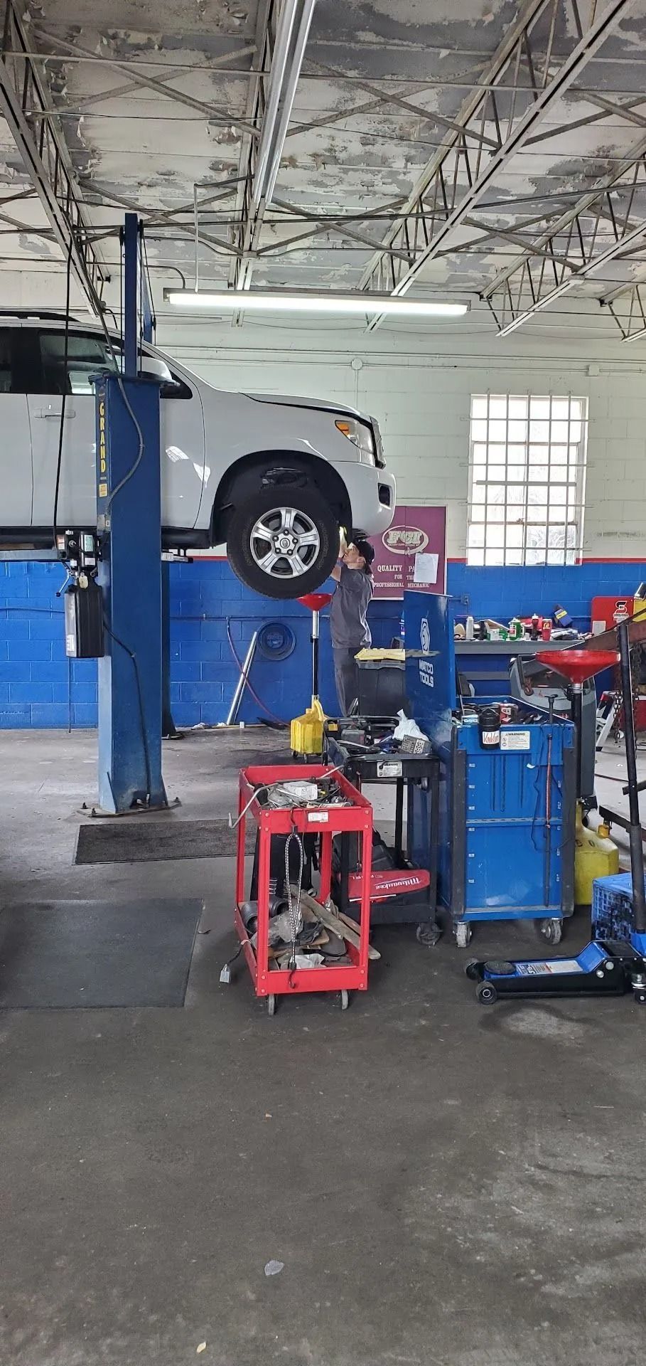 A white car raised on a lift in a garage. Mechanic working under the vehicle. Tools and equipment visible.