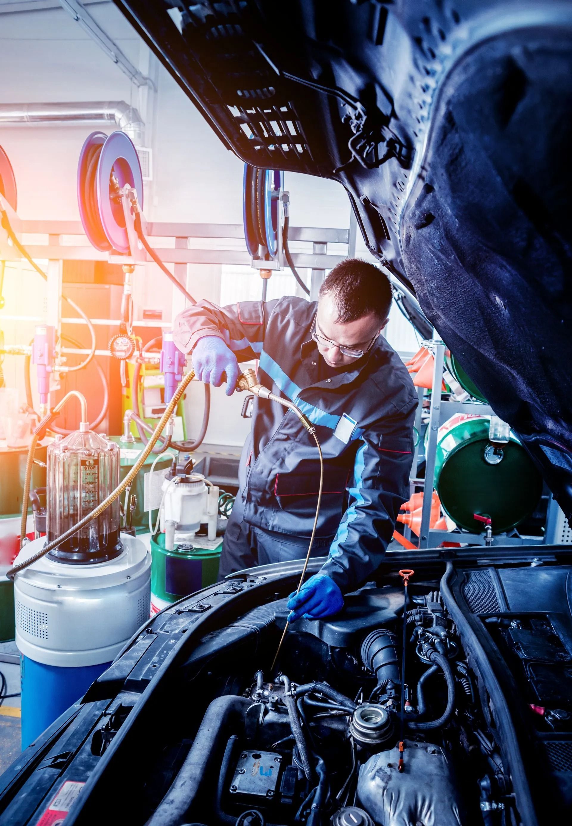 Mechanic in blue coveralls working on a car engine in a garage; using a hose.