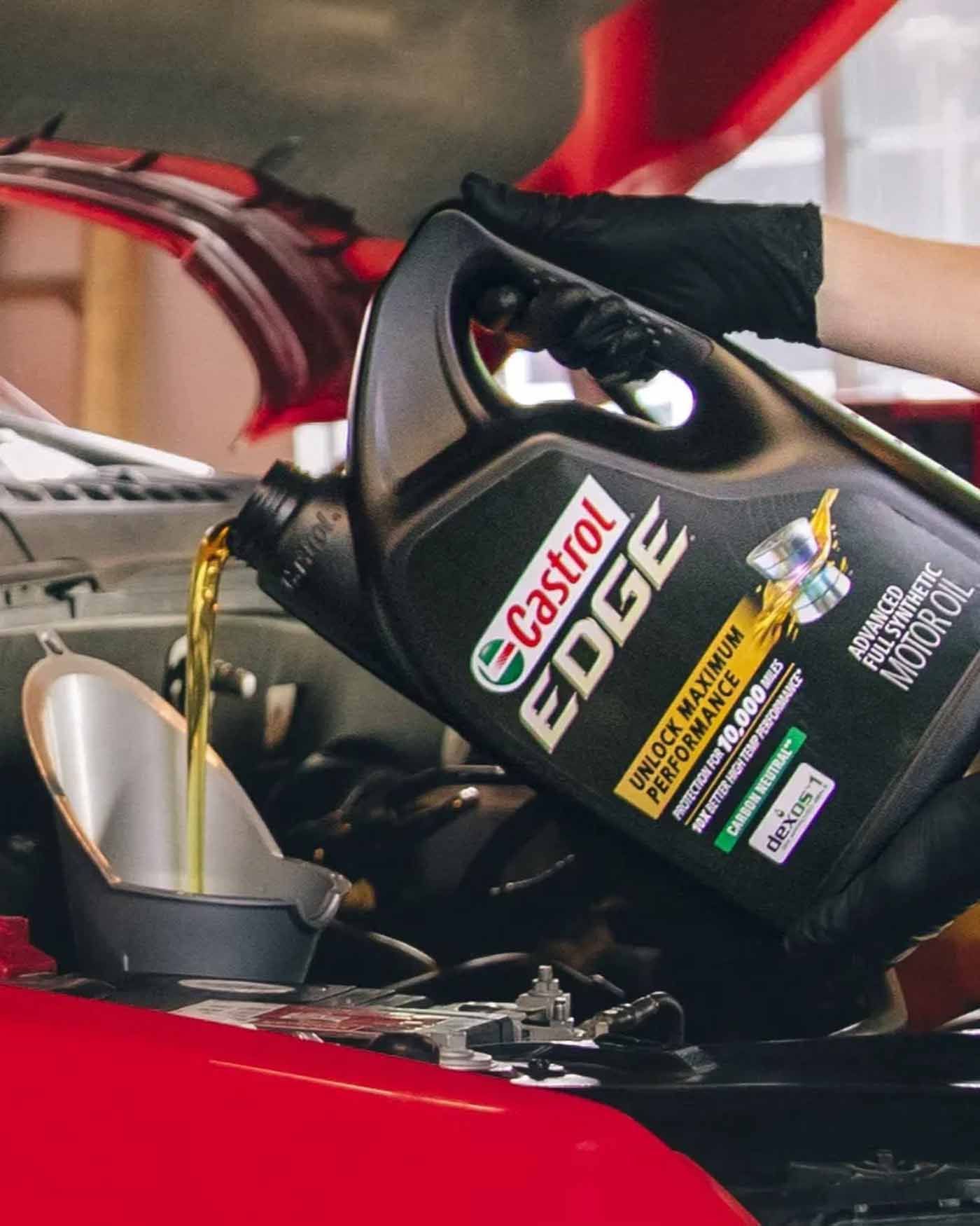 A person pours Castrol Edge motor oil into a car's engine. Black bottle and gloves, red car.