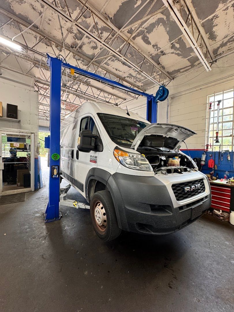 White Ram ProMaster van on a lift with the hood open in a garage, blue lift arms, tools.