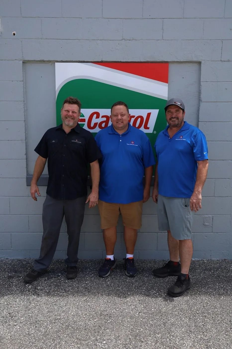 Three men pose in front of a Castrol sign on a gray building. Two wear blue shirts, one black.