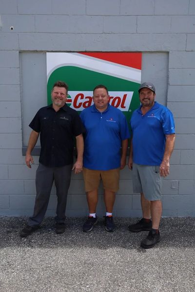 Three men pose in front of a Castrol sign on a gray building. Two wear blue shirts, one black.