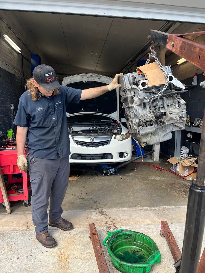Mechanic holding up a car engine in a garage with a car in the background and an oil pan.