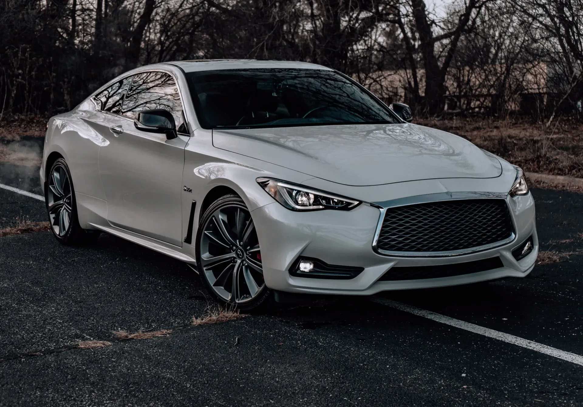 White Infiniti Q60 coupe on dark asphalt road, daytime, trees in background.