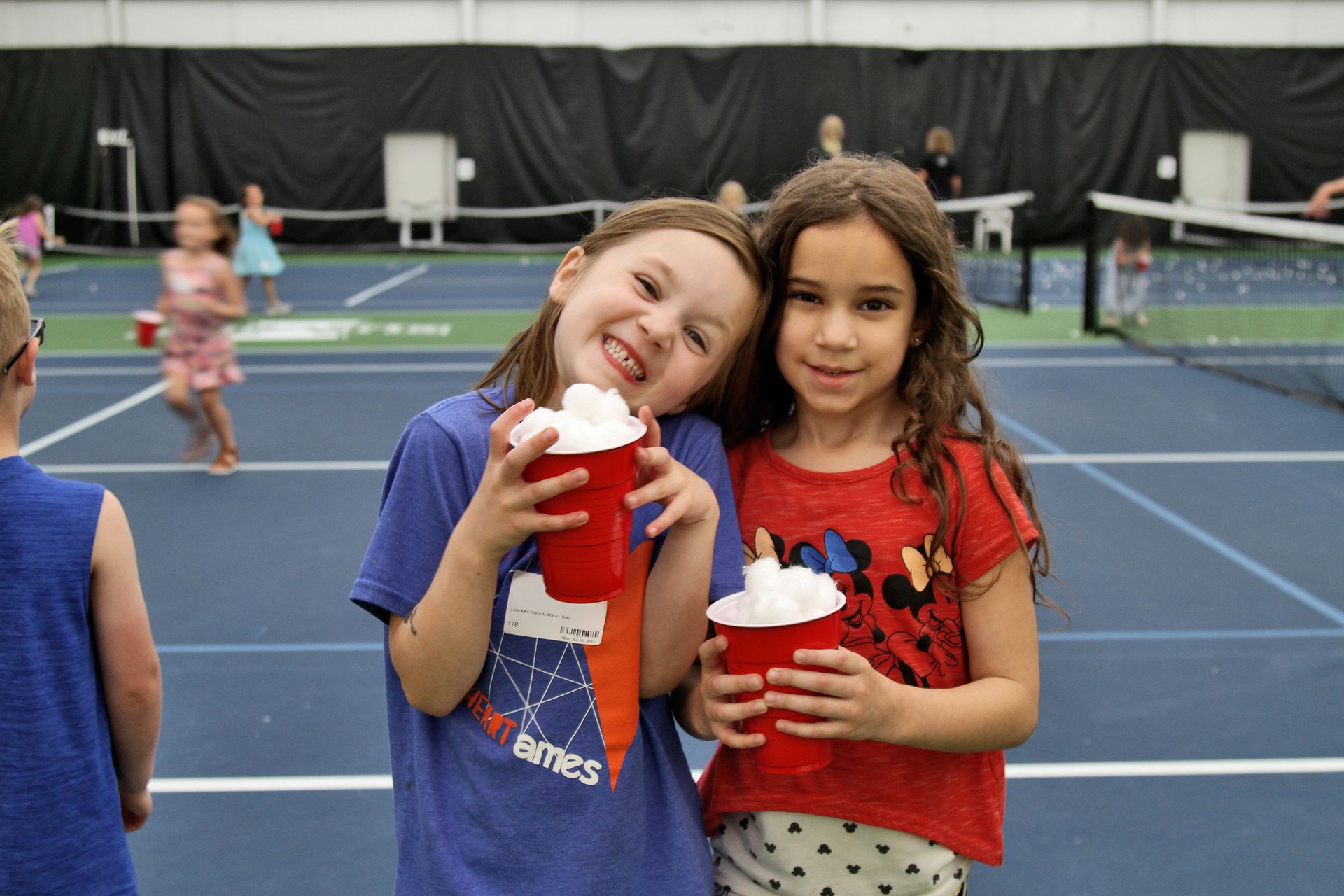 Two young girls are holding cups of hot chocolate on a tennis court.