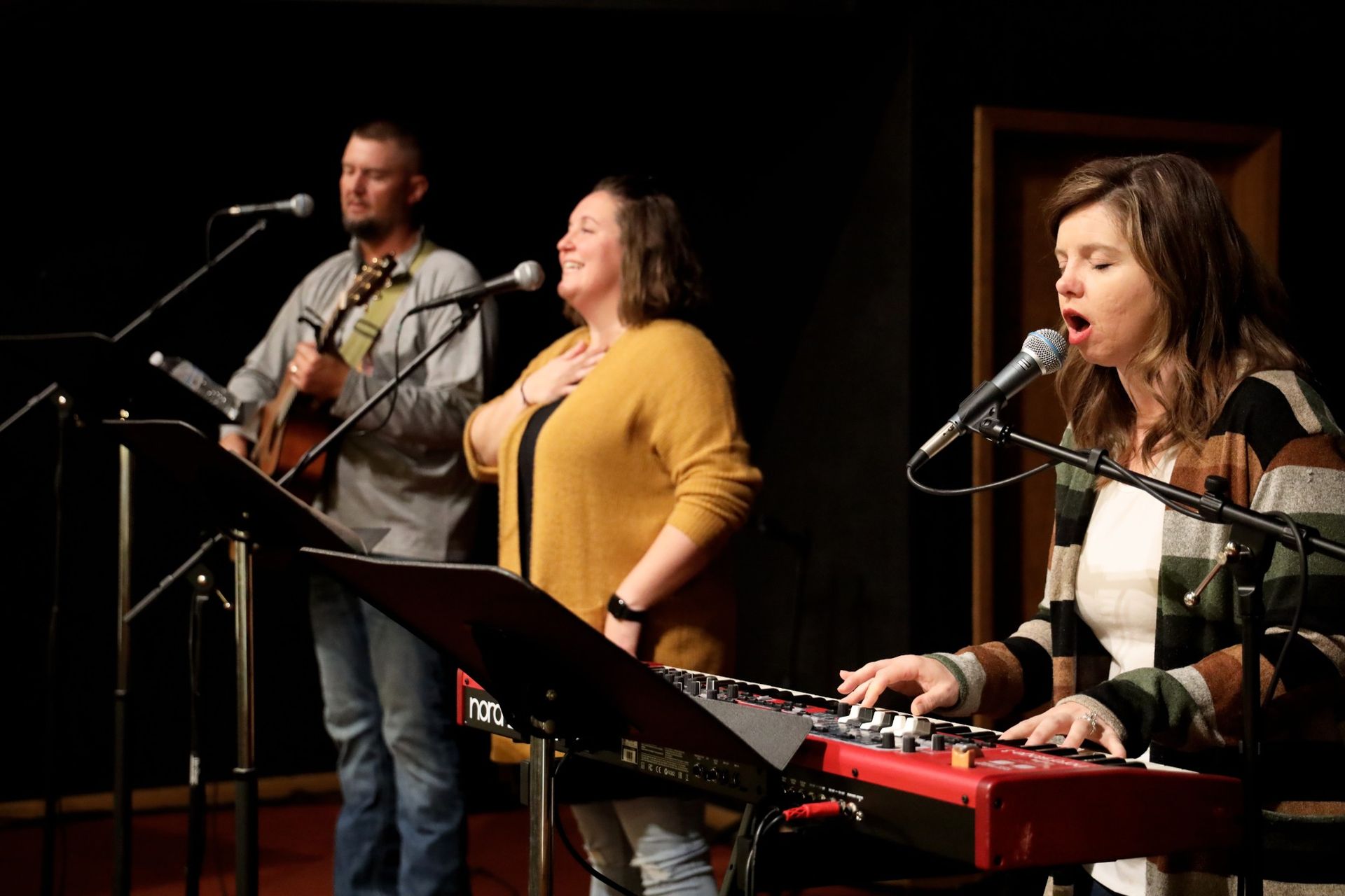 A musical group on stage: one playing keyboard, one singing, one with guitar and singing.