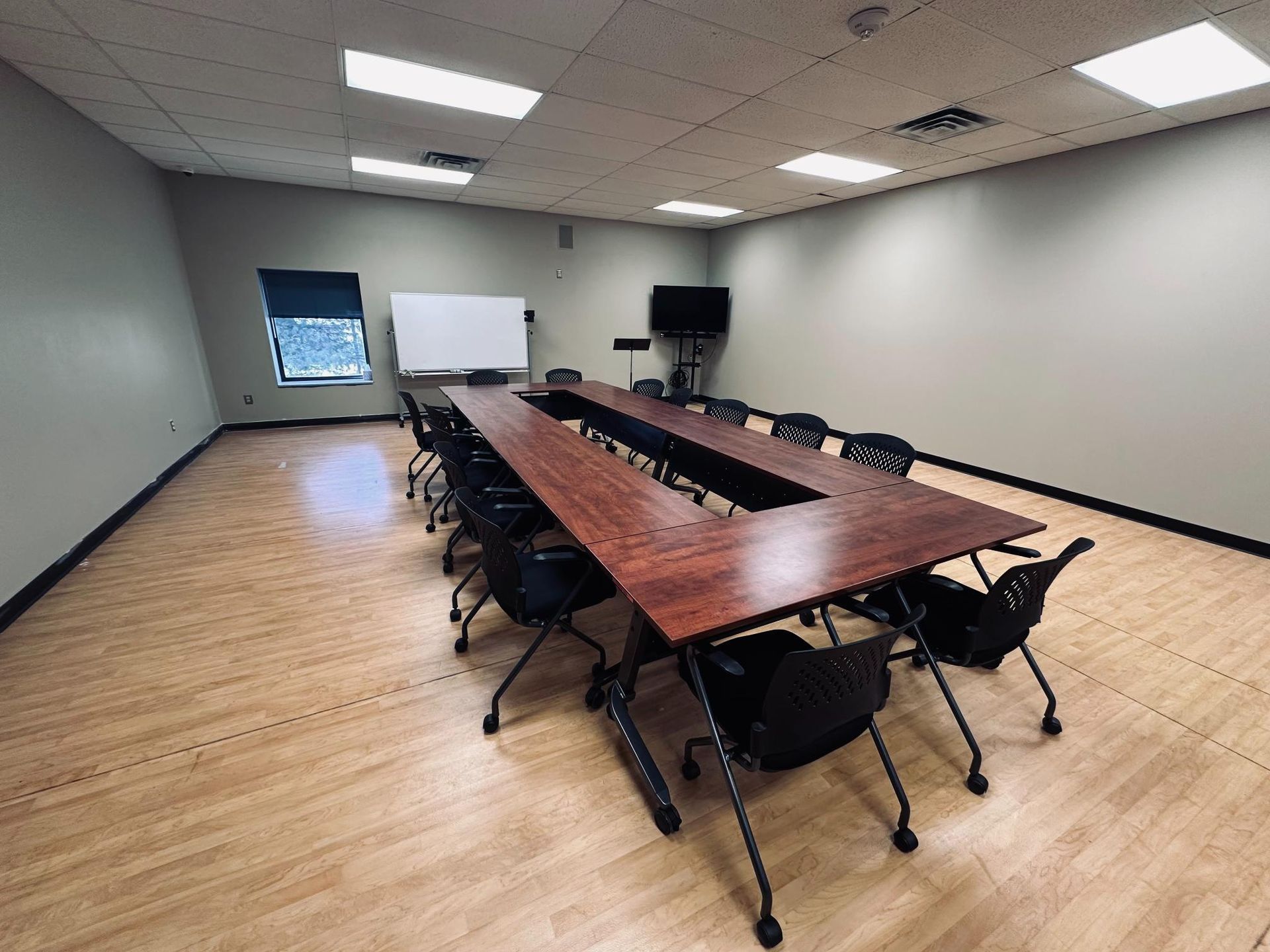 Conference room with a large wooden table surrounded by black chairs. Whiteboard and TV on wall.