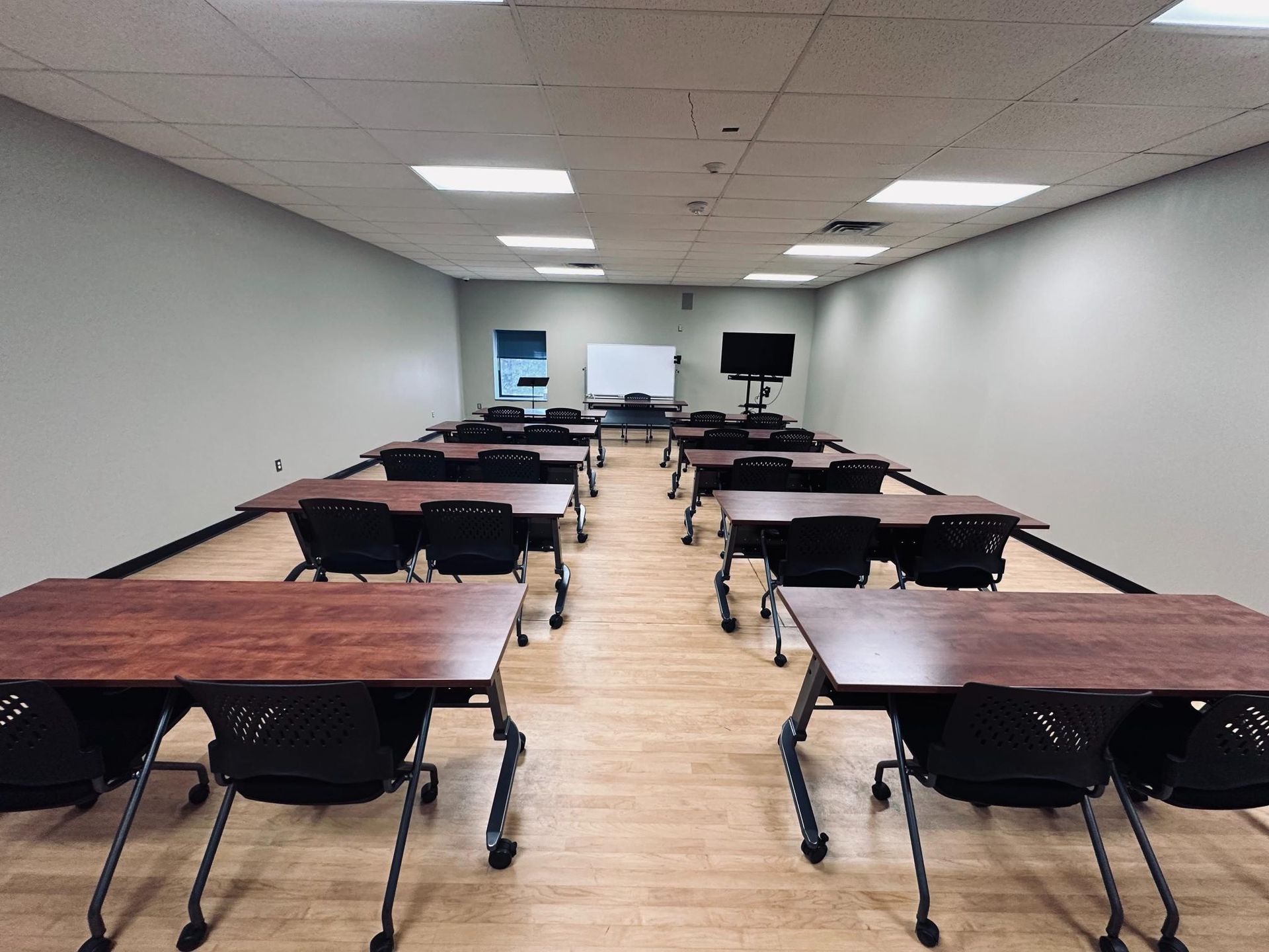 Classroom with rows of desks, a whiteboard, and a screen; beige walls, wooden floors, overhead lights.