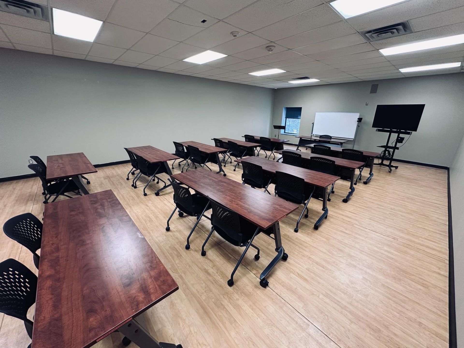 Empty classroom with rows of tables and chairs, whiteboard, and TV on a stand.