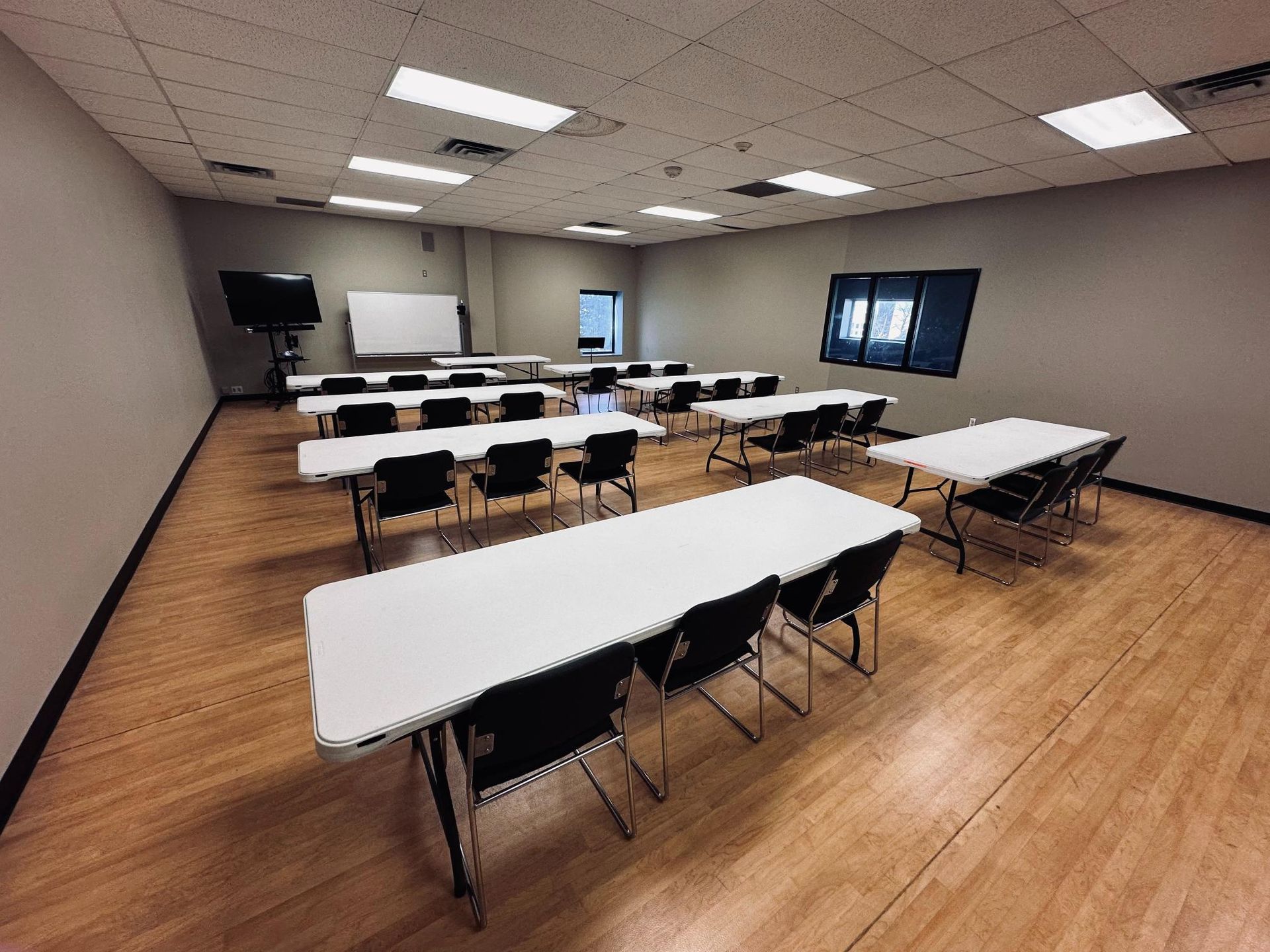 Classroom with rows of white tables and black chairs, whiteboard, and multiple screens.