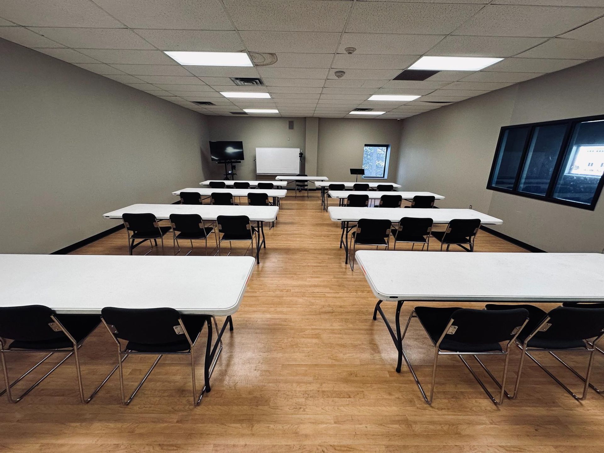 Classroom with rows of white tables, black chairs, whiteboard, and rectangular windows.