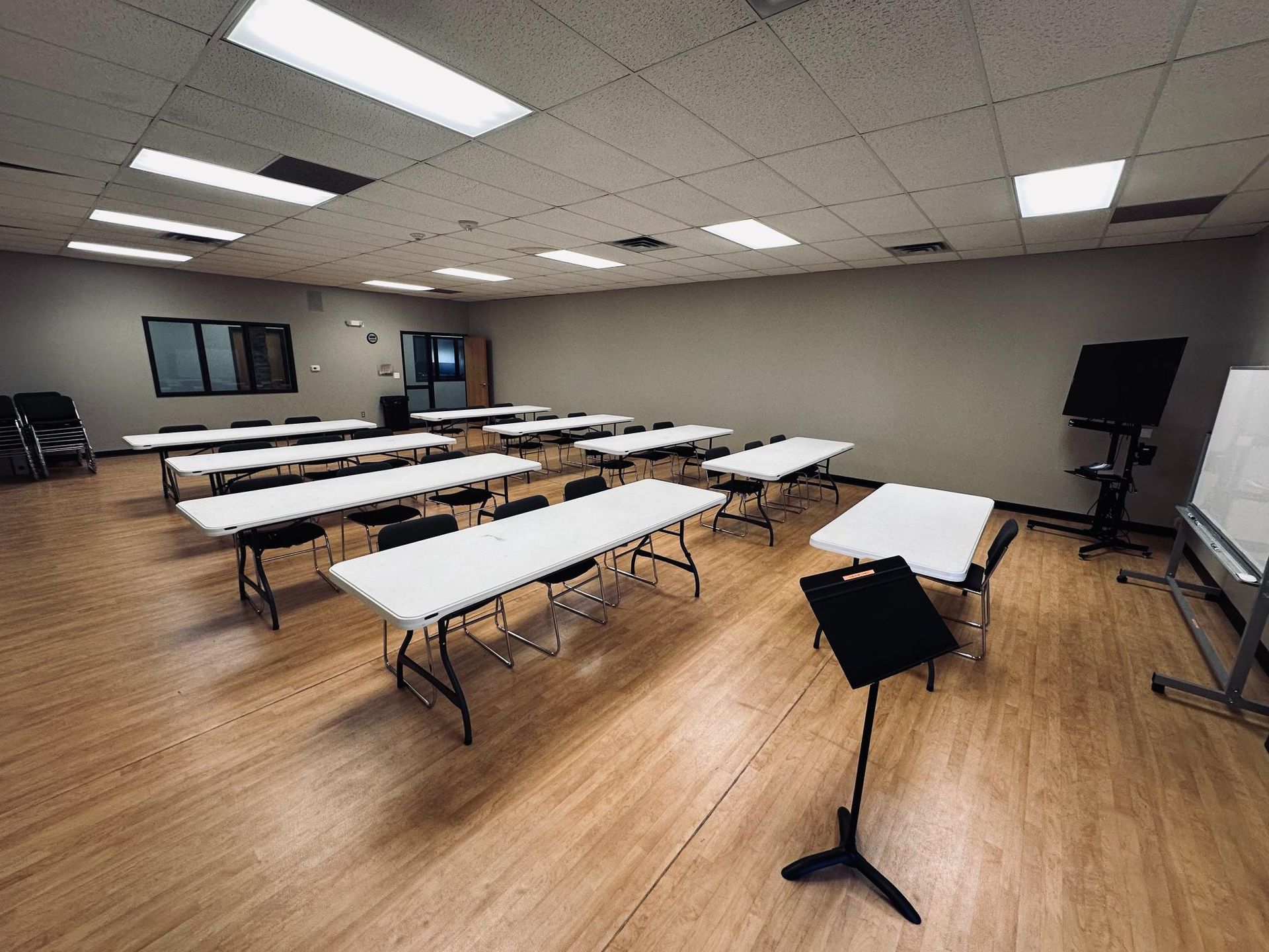 Empty classroom with rows of tables, chairs, and musical stand. Wooden floor, neutral walls, and overhead lights.