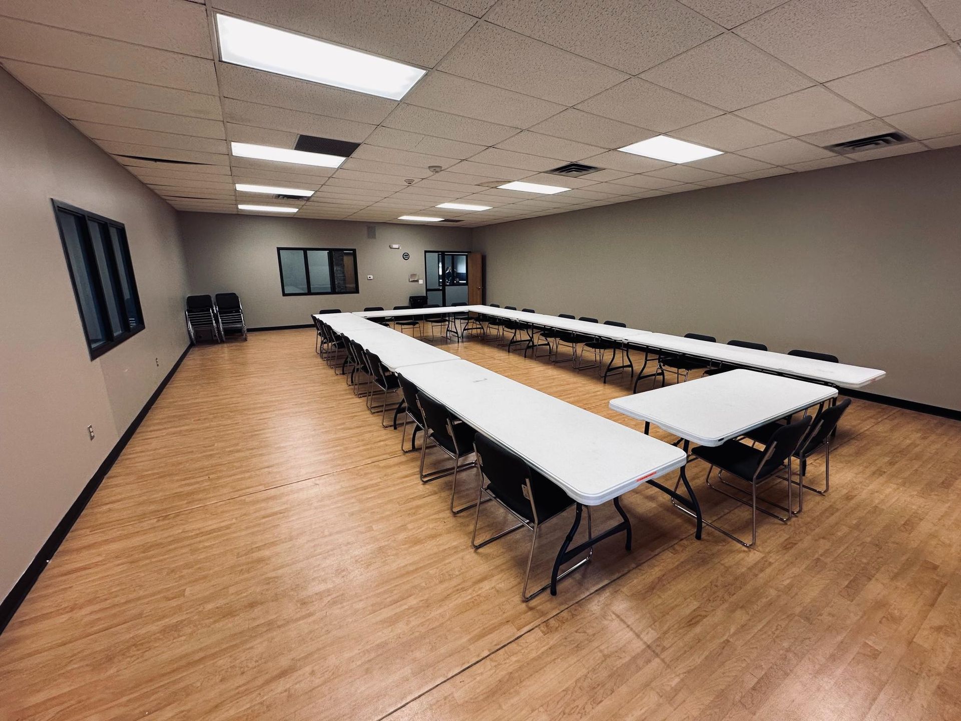 Empty conference room with tables arranged in a U-shape, chairs, and wood-look flooring.