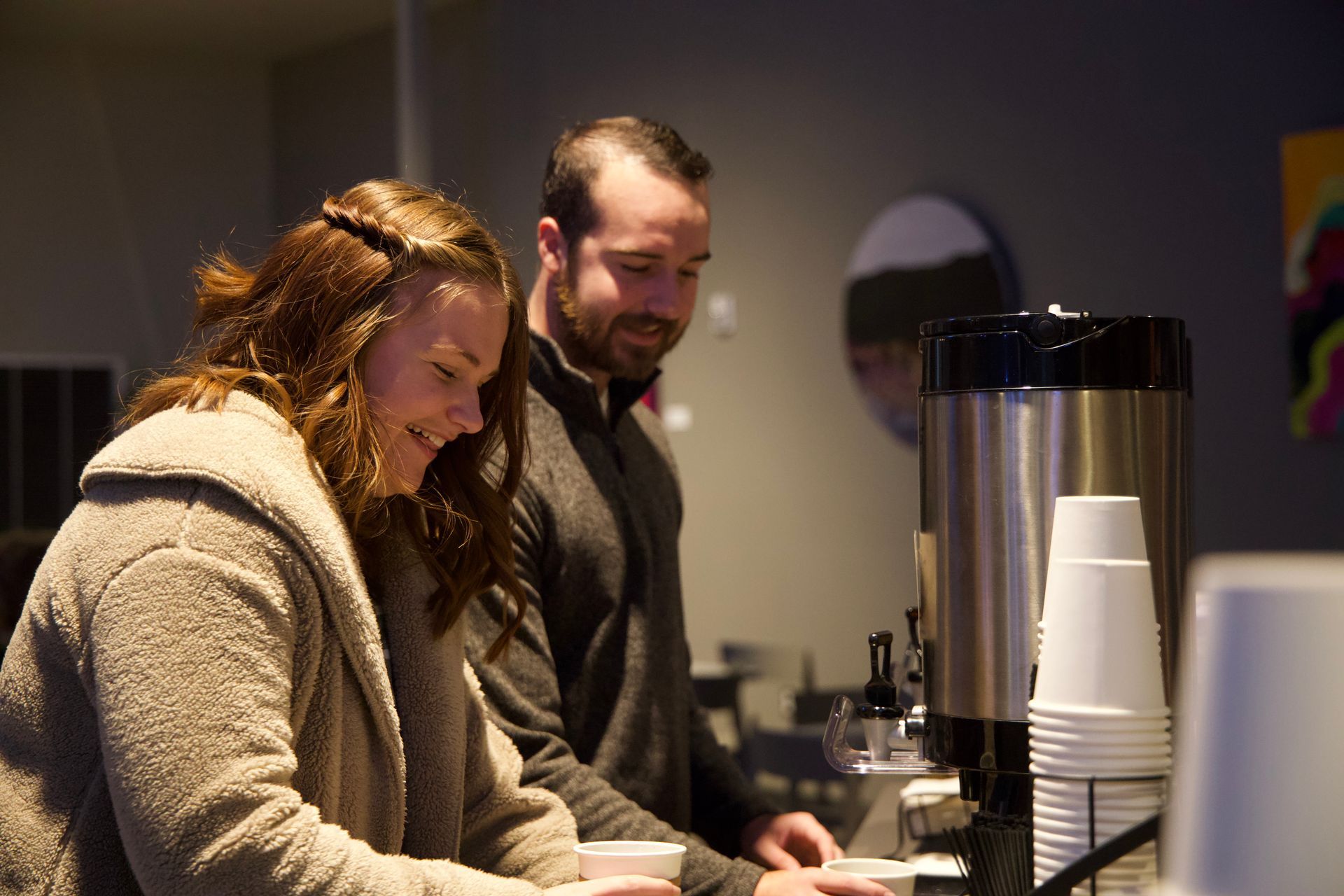 Woman and man pouring coffee, smiling. Light-colored coats, coffee station in a room.