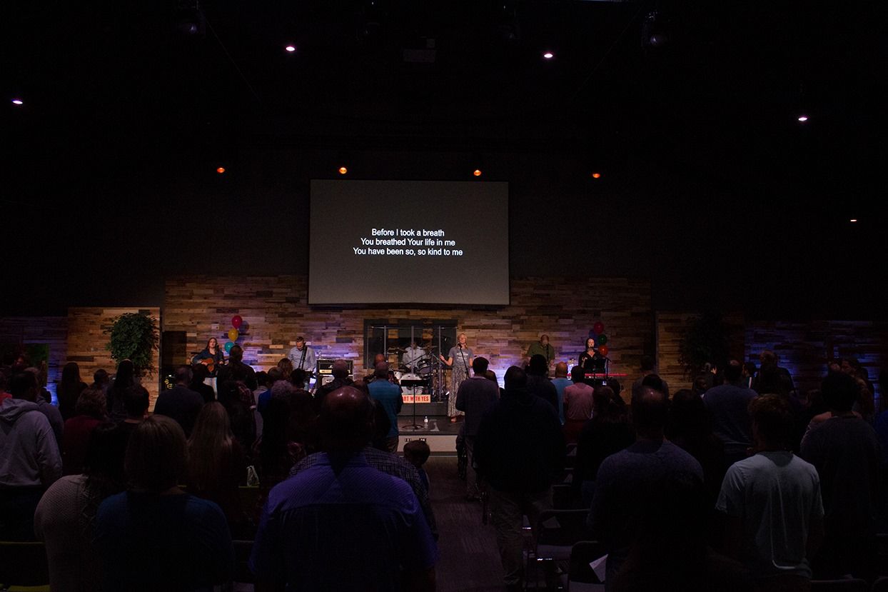 People in a church service with a band on stage, singing along with lyrics on a screen.