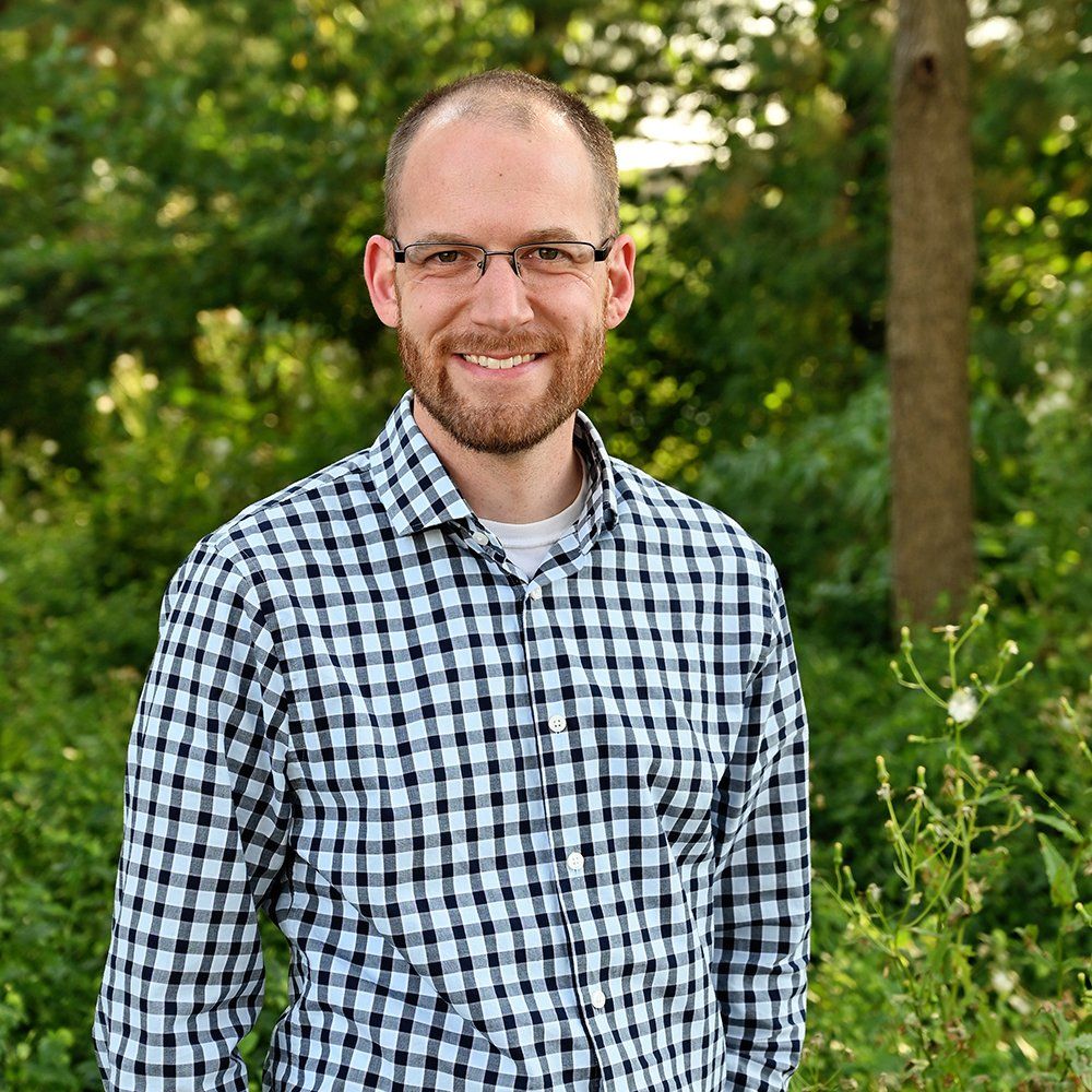 A man wearing glasses and a plaid shirt is standing in front of a forest.