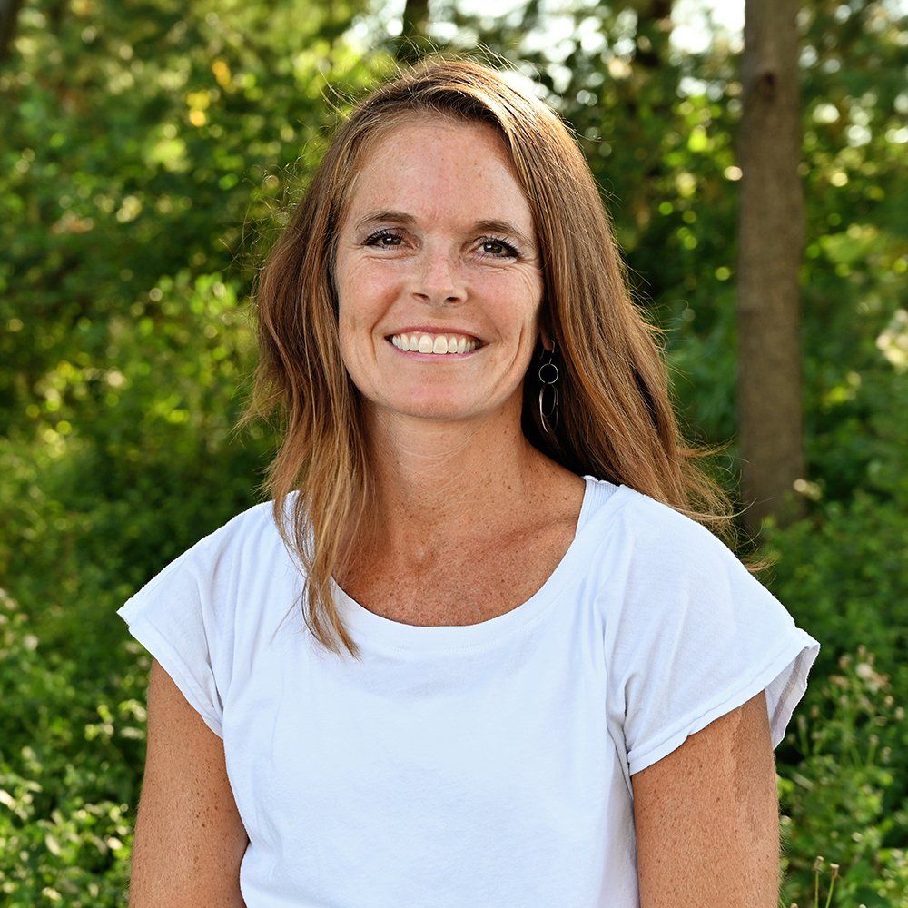 A woman in a white shirt is smiling in front of trees.