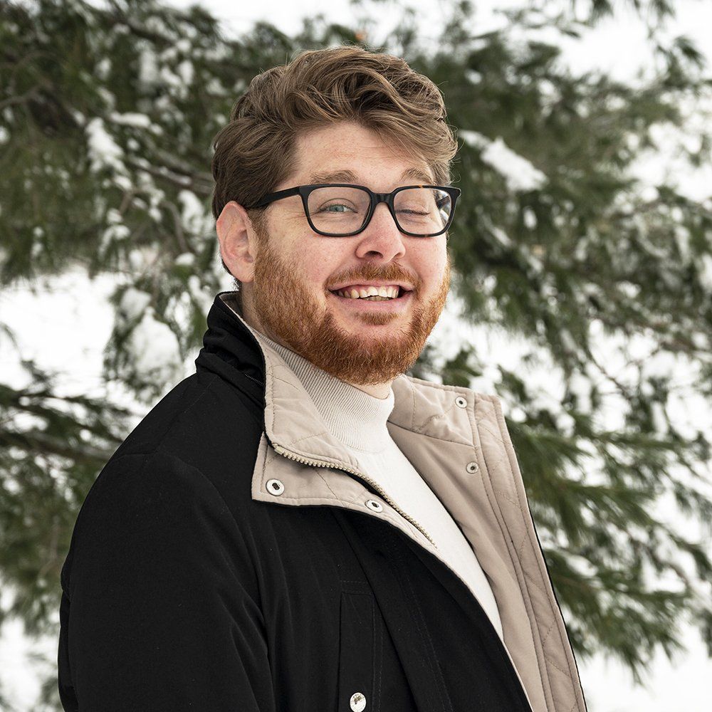 A man with a beard and glasses is smiling in front of a tree.