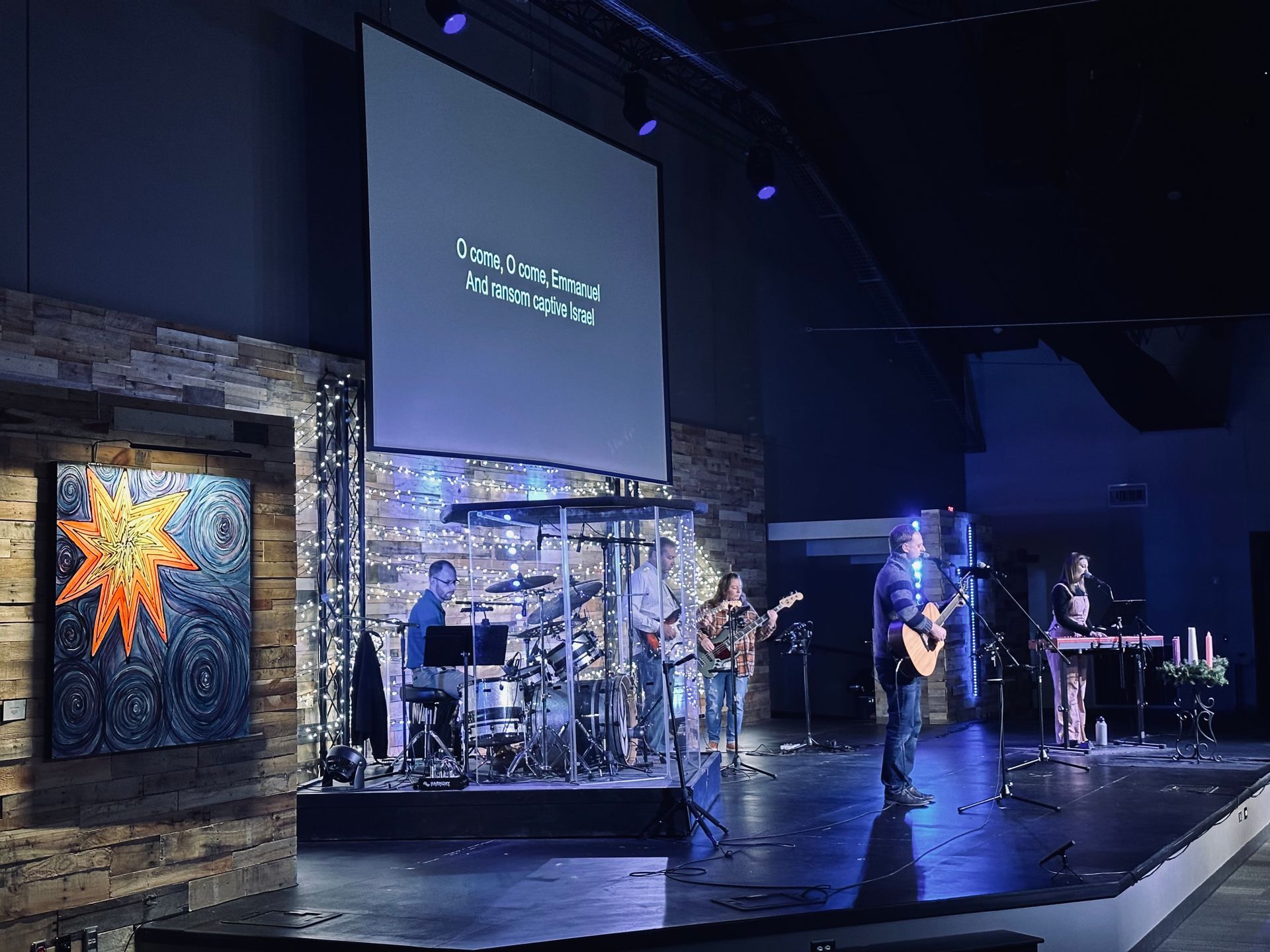 A band performs on stage in a church. A screen displays lyrics, blue lighting illuminates the stage.