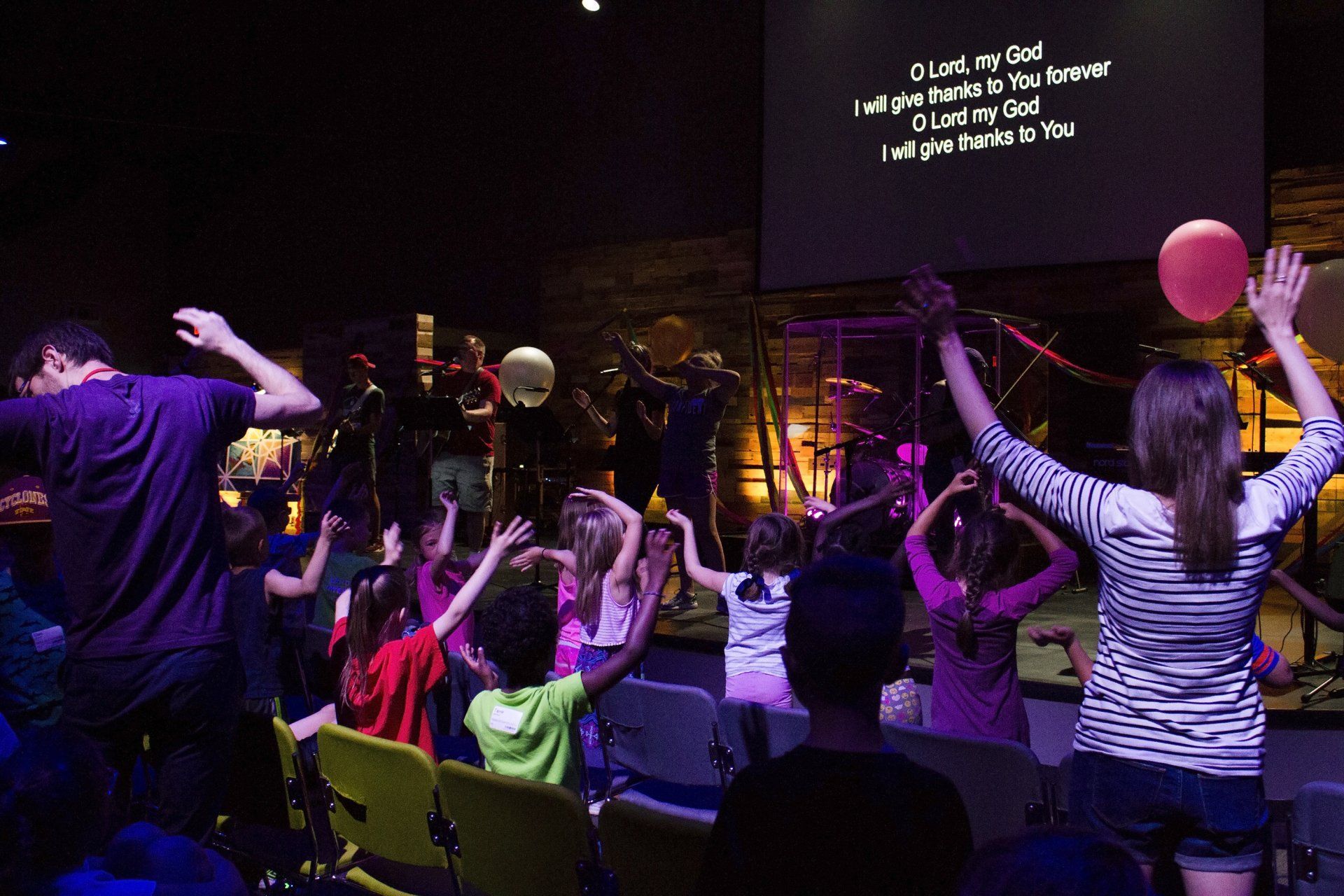 People with raised hands worshiping in a dimly lit room with a screen displaying lyrics.