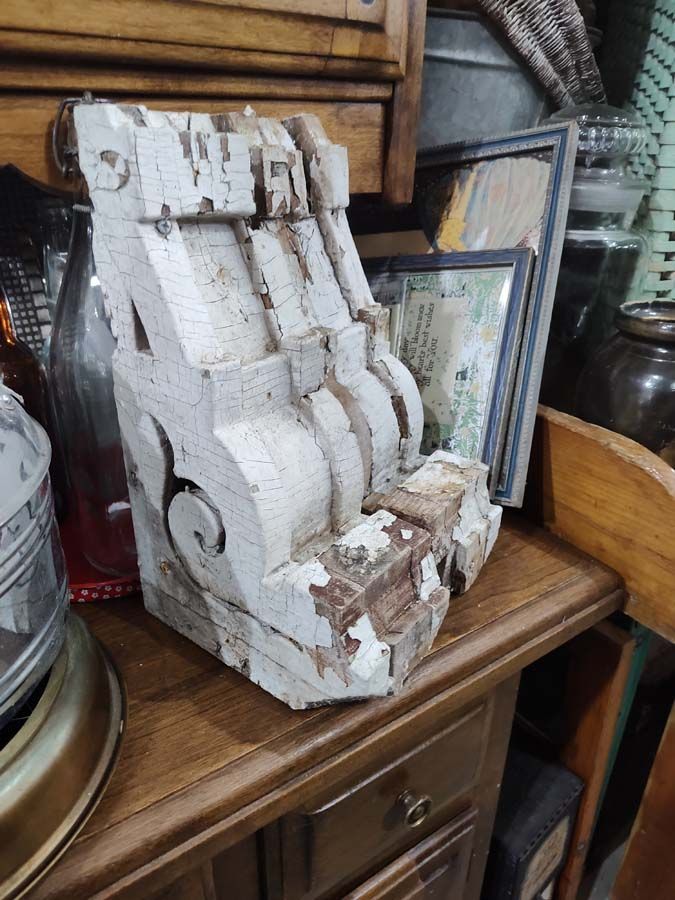 Weathered, white-painted wooden architectural bracket with decorative scrollwork, displayed on a wooden cabinet.