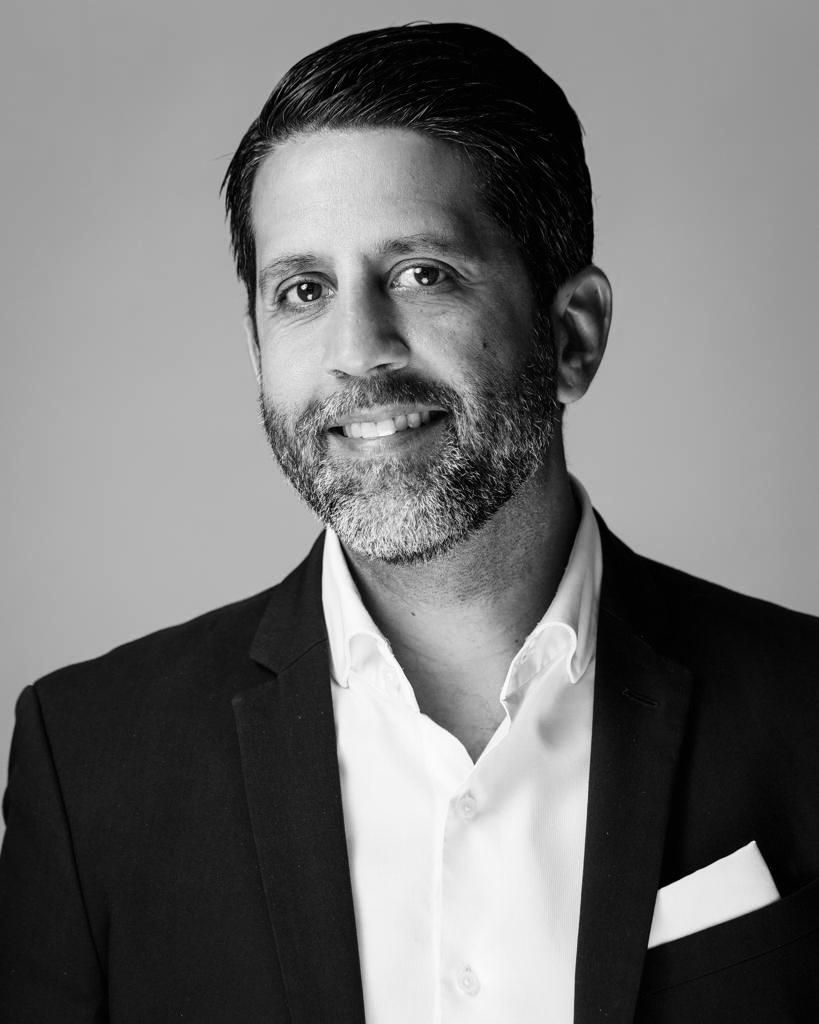 Studio portrait of a smiling man in a dark suit and white shirt against a gray background