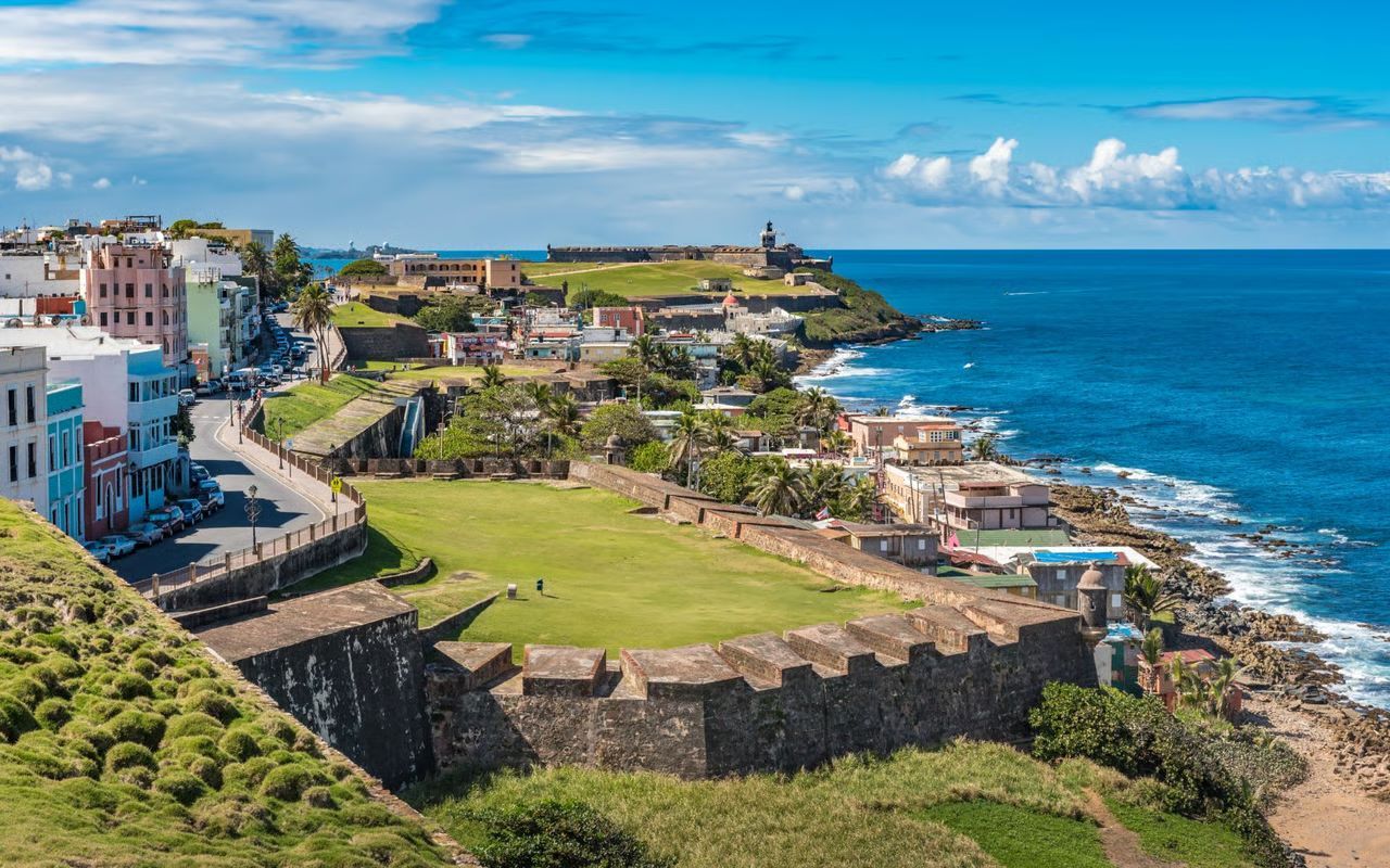 Colorful buildings line a coast under a bright blue sky, with ocean waves breaking in the foreground.