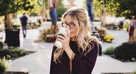 Woman with glasses drinking from a white cup on a sunny outdoor walkway with blurred flowers and people in the background