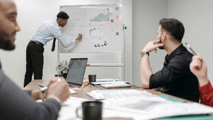 Team meeting in a conference room, with one person presenting at a whiteboard and others listening at a table.