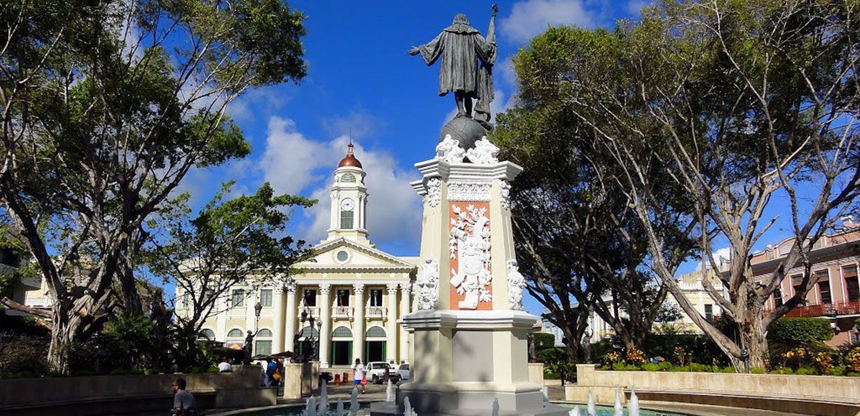 Statue of a man in front of a church, trees frame the view. Bright blue sky.